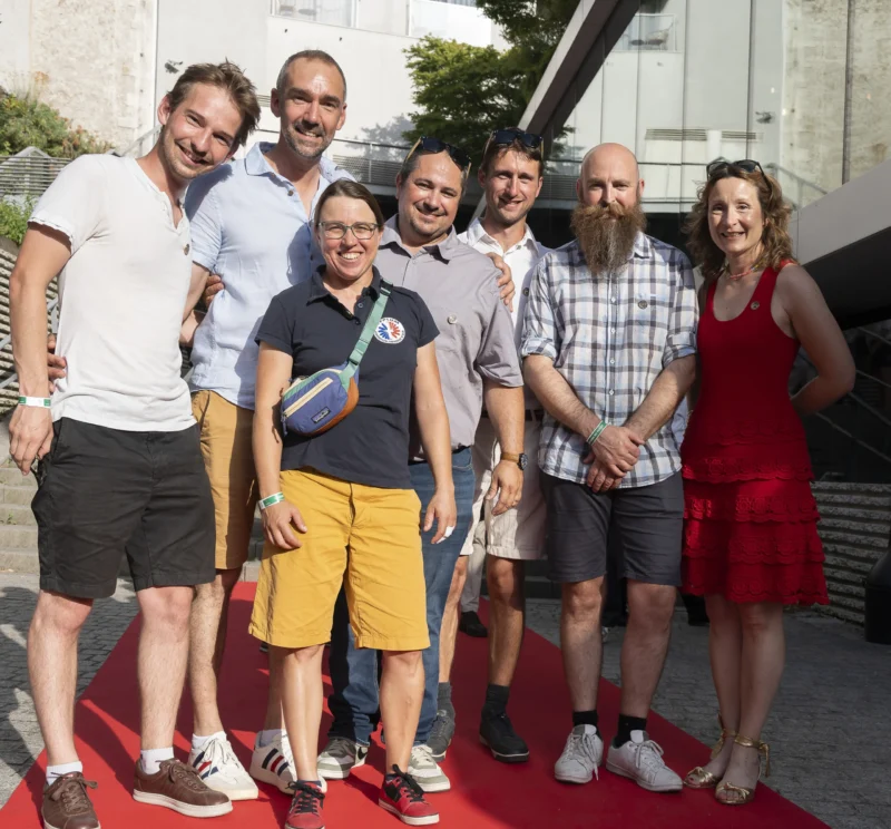 Cette photographie montre un groupe de sept personnes posant ensemble sur un tapis rouge. Le sujet principal est leur rassemblement joyeux, avec une personne tenant un ballon de rugby.La composition est symétrique, avec les individus alignés. Le cadrage est serré, mettant en valeur les visages et les expressions. La lumière est naturelle et douce, probablement en extérieur. Les couleurs dominantes sont vives : rouge du tapis, tons pastel et neutres des vêtements.Le style est informel et décontracté, évoquant une ambiance conviviale et festive.