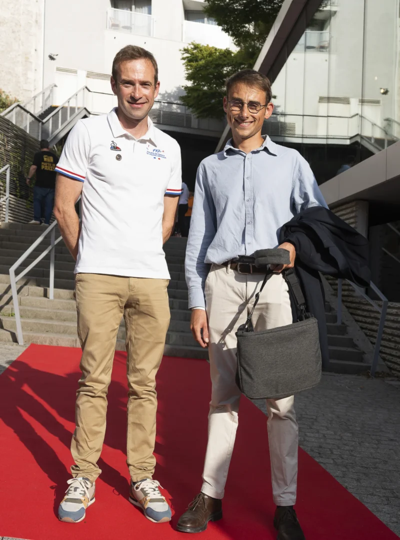 Cette photographie montre deux hommes posant sur un tapis rouge. Le sujet principal est ces deux individus, l’un vêtu d’un polo blanc et d’un pantalon beige, l’autre en chemise bleue et pantalon clair avec une veste beige.La composition est symétrique, avec un cadrage serré sur les deux hommes. L’arrière-plan inclut des structures architecturales et un escalier. La lumière est naturelle et douce, suggérant un jour ensoleillé. Les couleurs dominantes sont les tons pastel (bleu, beige) et le rouge vif du tapis.Le style est sobre et professionnel, évoquant une ambiance formelle, probablement liée à un événement public ou une cérémonie.