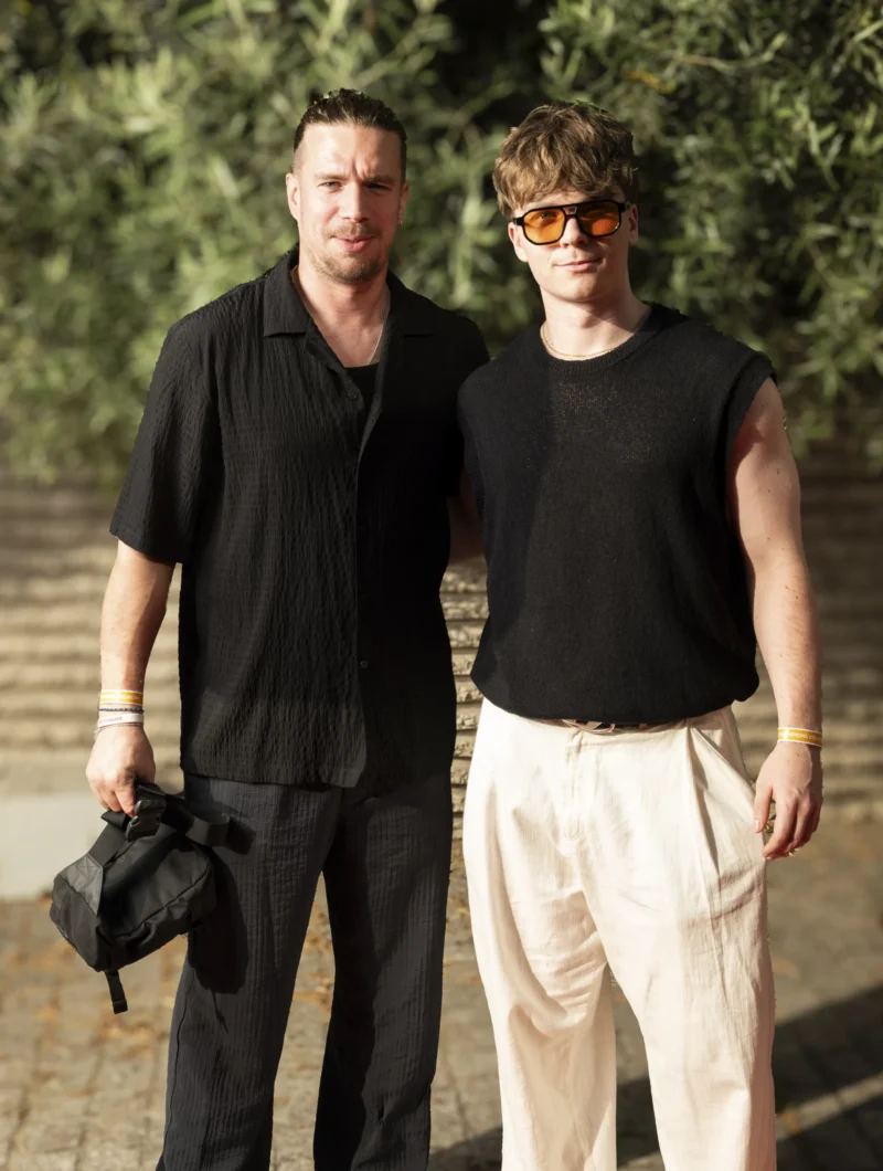 Cette photographie met en scène deux hommes posant côte à côte. Le sujet principal est leur apparence décontractée et stylisée.La composition est symétrique, avec un cadrage serré sur les deux individus. La lumière semble naturelle, douce et légèrement tamisée, créant des ombres subtiles. Les couleurs dominantes sont les tons neutres : noir (vêtements), blanc (pantalon), et des touches de vert en arrière-plan grâce à la végétation.Le style est urbain et minimaliste, évoquant une ambiance décontractée et moderne. L'atmosphère est calme et élégante.