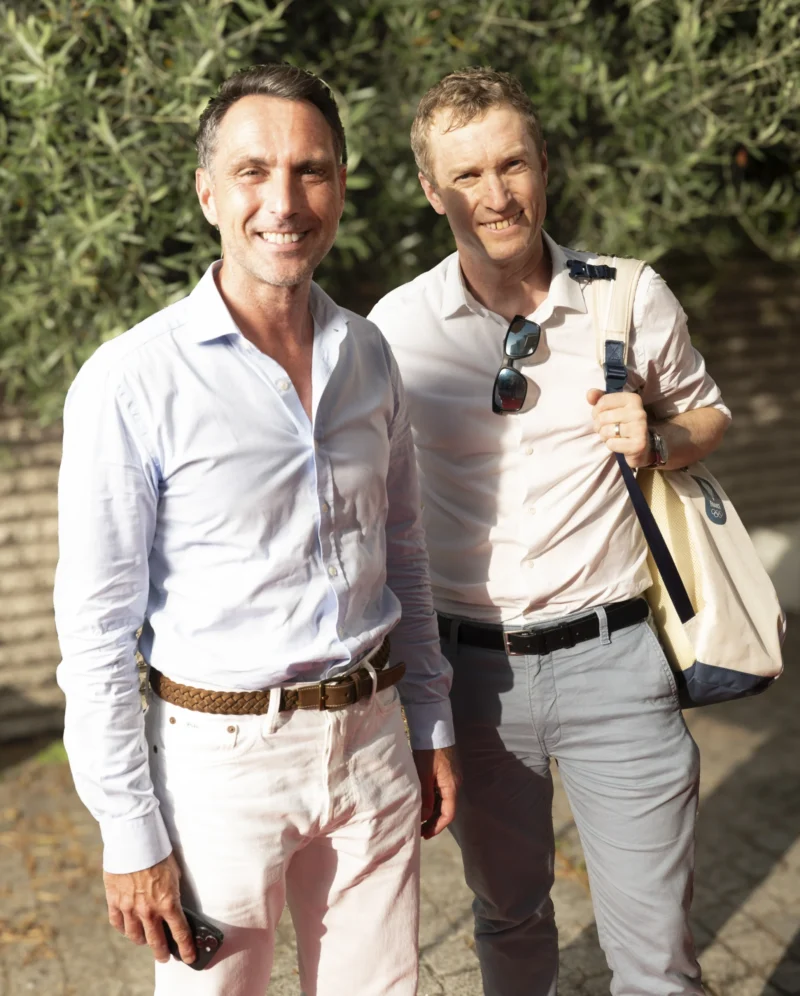 Cette photographie montre deux hommes souriants posant côte à côte. Le sujet principal est leur complicité et leur apparence décontractée.La composition est symétrique, avec un cadrage serré sur les épaules et le haut du corps. La lumière semble naturelle, douce et légèrement tamisée, créant des ombres subtiles. Les couleurs dominantes sont des tons pastel : bleu clair, blanc et beige, renforçant une ambiance détendue et estivale. Le style est informel et rétro, évoquant une ambiance chaleureuse et conviviale des années 1980 ou 1990.