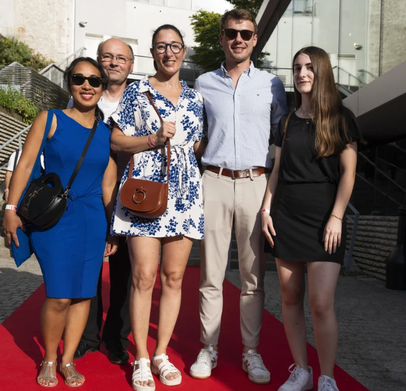 Cette photographie montre cinq personnes posant sur un tapis rouge. Le sujet principal est le groupe, composé de quatre adultes et un aîné, souriants et habillés élégamment.La composition est symétrique, avec les individus alignés côte à côte. Le cadrage est serré, mettant l’accent sur les visages et les tenues. La lumière est naturelle et douce, probablement en extérieur en journée. Les couleurs dominantes sont vives : bleu, noir, blanc et motifs floraux.Le style est décontracté mais soigné, évoquant une ambiance festive ou un événement public. L’ambiance est chaleureuse et conviviale.