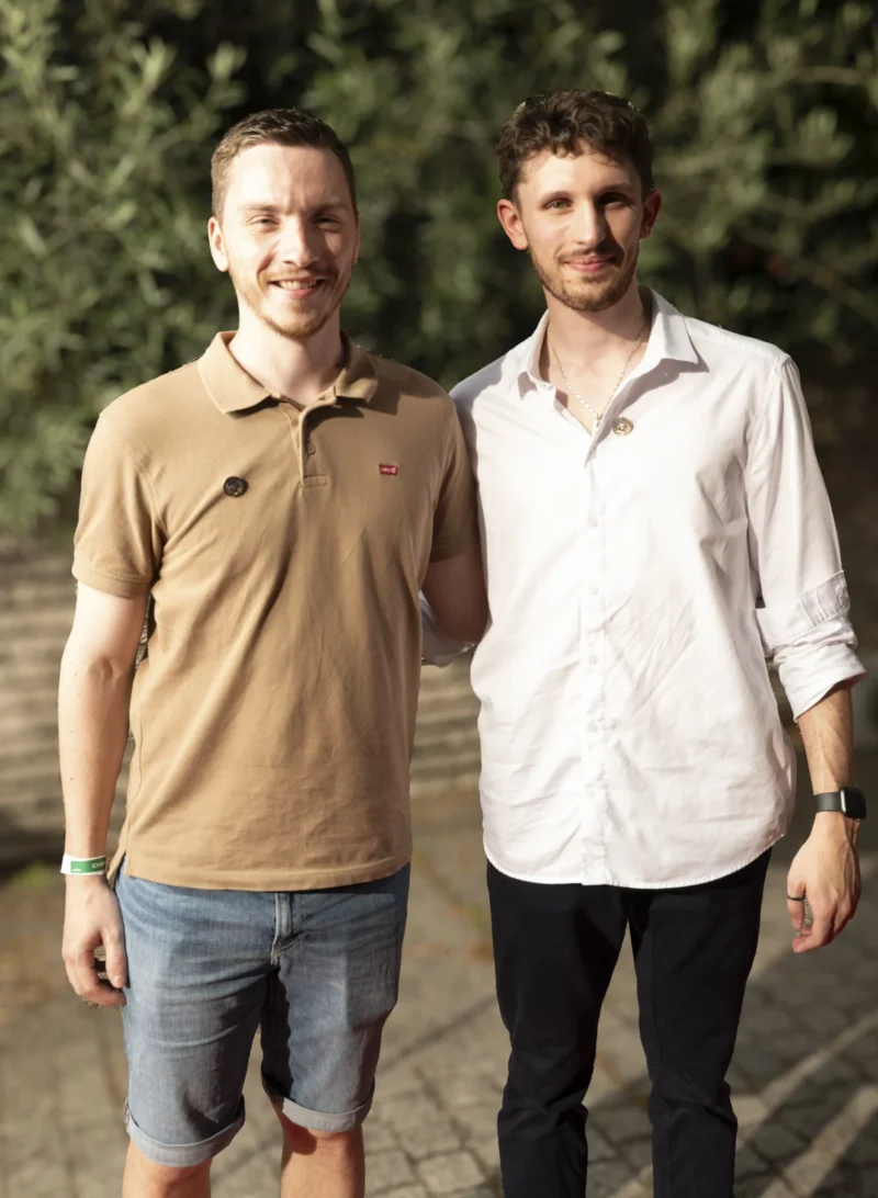 Cette photographie montre deux hommes posant côte à côte. Le sujet principal est leur présence conjointe, suggérant une relation amicale ou professionnelle.La composition est symétrique, avec un cadrage serré sur les épaules et le haut du corps. La lumière est naturelle et douce, probablement en extérieur, créant des ombres légères. Les couleurs dominantes sont neutres : beige, blanc et bleu clair, avec des touches de vert en arrière-plan.Le style est simple et décontracté, évoquant une ambiance détendue et informelle.