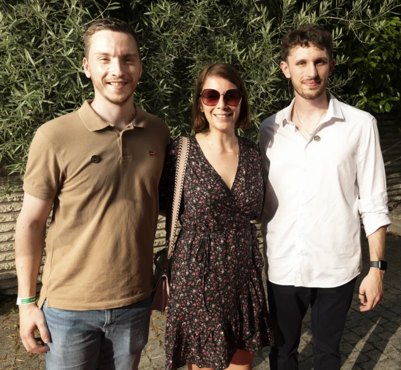 Cette photographie montre trois personnes posant côte à côte devant un fond de végétation. Le sujet principal est ce trio souriant, formant une composition symétrique et harmonieuse.Le cadrage est serré sur les épaules et le haut du corps. La lumière est naturelle et douce, probablement en extérieur. Les couleurs dominantes sont des tons neutres et naturels : vert pour le fond, marron et blanc pour les vêtements des hommes, et des teintes florales pour la robe de la femme.L’ambiance est détendue et conviviale, évoquant un moment informel et joyeux.