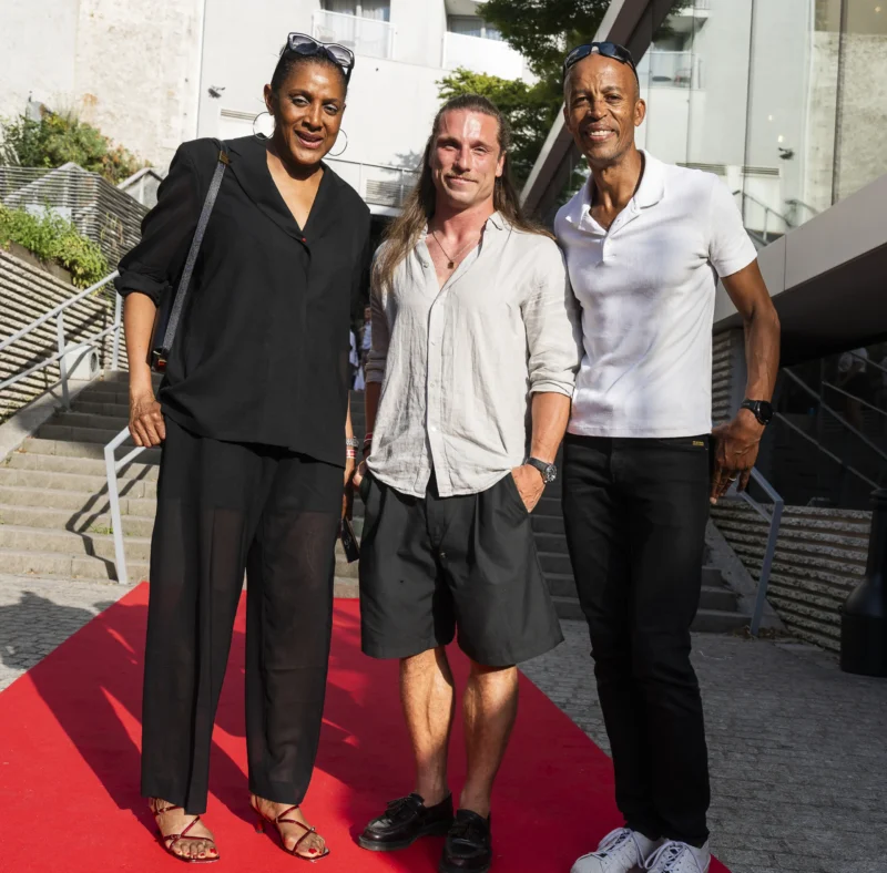 Cette photographie montre trois personnes posant sur un tapis rouge. Le sujet principal est leur interaction souriante, suggérant un événement formel.La composition est symétrique, avec les trois individus alignés verticalement. Le cadrage est serré, mettant en valeur leurs visages et tenues. La lumière est naturelle et douce, probablement en extérieur en journée.Les couleurs dominantes sont les tons neutres (noir, blanc, gris) et les touches de rouge du tapis. Le style est élégant et décontracté à la fois. L’ambiance est conviviale et professionnelle, typique d’un événement mondain.