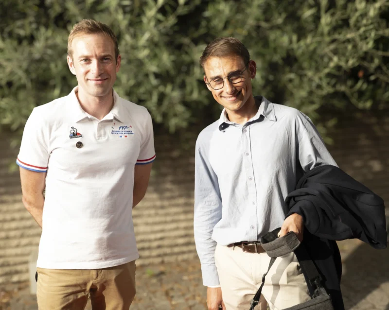 Cette photographie montre deux hommes souriants posant côte à côte. Le sujet principal est leur interaction amicale.La composition est symétrique, avec un cadrage serré sur les épaules et le haut du corps. L’arrière-plan flou met en valeur des arbres et une lumière naturelle douce. Les couleurs dominantes sont les tons pastel (bleu clair, blanc) et les verts naturels.Le style est simple et spontané, évoquant une ambiance détendue et chaleureuse, probablement prise en extérieur lors d’une journée ensoleillée.