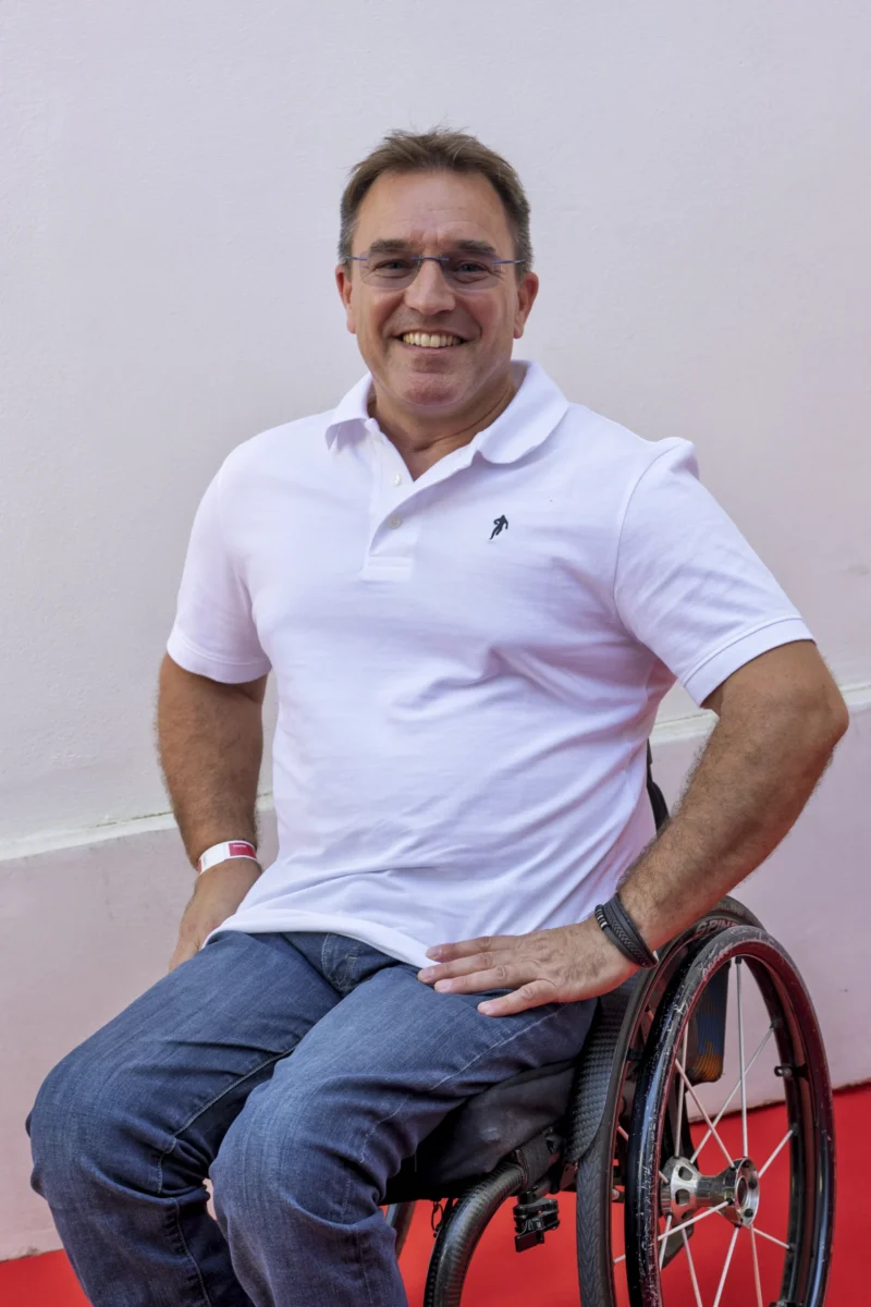 Cette photographie met en scène un homme souriant assis dans un fauteuil roulant, vêtu d’un polo blanc et d’un jean. Le cadrage est serré, centré sur lui, avec un fond neutre et légèrement coloré.La lumière est douce et naturelle, éclairant son visage et créant une ambiance chaleureuse. Les couleurs dominantes sont les tons pastel (blanc, bleu clair) et les touches de rouge du fauteuil. Le style est simple et direct, mettant en valeur la positivité et la sérénité du sujet. L’ambiance est accueillante et humaine.