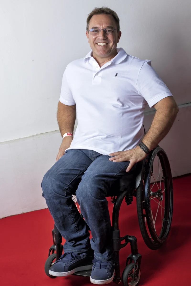 Cette photographie met en scène un homme souriant assis dans un fauteuil roulant, qui semble être le sujet principal. Le cadrage est serré, centré sur lui, avec un fond neutre et clair.La lumière est douce et naturelle, éclairant uniformément son visage et créant une ambiance chaleureuse. Les couleurs dominantes sont les tons pastel : blanc (chemise), bleu clair (pantalon) et rouge (sol), renforçant une ambiance positive et apaisante. Le style est simple et direct, mettant l'accent sur l'humanité et la sérénité.