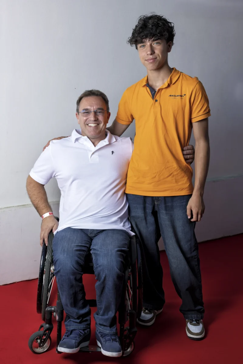 Cette photographie met en scène deux hommes en position debout. Le sujet principal semble être leur relation, avec un homme assis en fauteuil roulant et un autre debout à ses côtés, posant une main sur son épaule.La composition est simple et symétrique, avec un cadrage serré sur les deux individus. La lumière est douce et naturelle, éclairant uniformément les visages. Les couleurs dominantes sont neutres (tons de gris du mur) avec des touches vives : le blanc et bleu du polo du premier homme, et l’orange du polo du second.L’ambiance est chaleureuse et humaine, évoquant complicité et soutien.