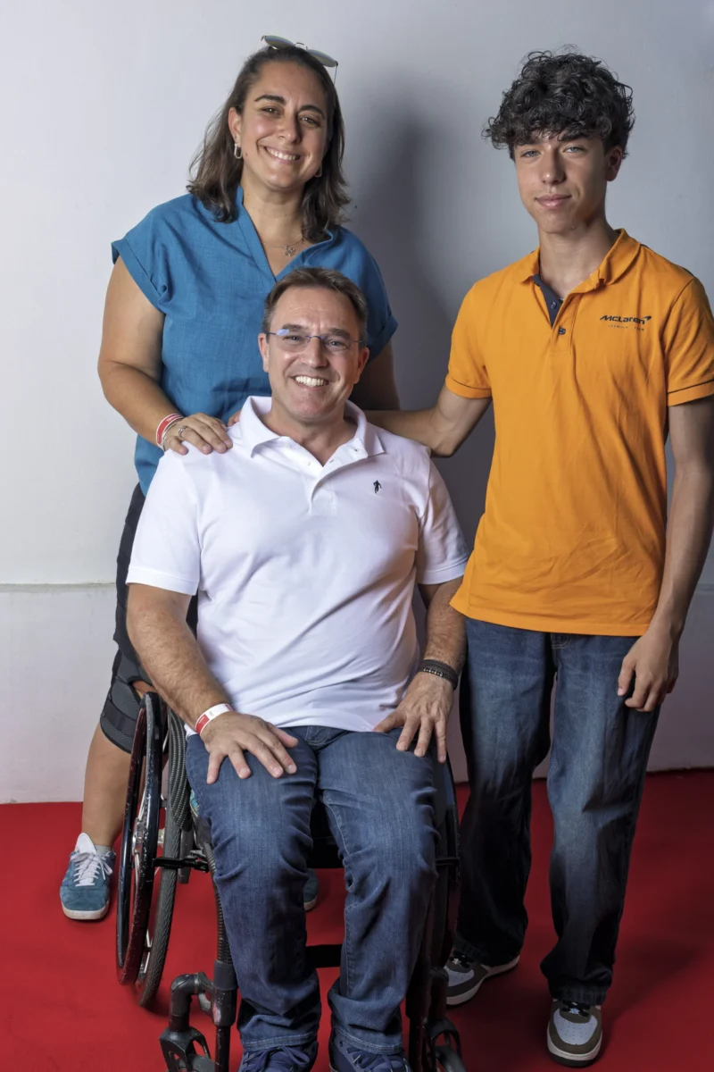 Cette photographie met en scène trois personnes souriantes. Le sujet principal est l’homme assis en fauteuil roulant, entouré de deux jeunes adultes, probablement des proches. La composition est symétrique et harmonieuse, avec un cadrage serré sur les trois individus.La lumière est douce et naturelle, éclairant uniformément les visages. Les couleurs dominantes sont vives : bleu, orange et blanc, créant une ambiance chaleureuse et positive. Le style est simple et direct, évoquant une scène authentique et familiale. L’ambiance est conviviale et rassurante.
