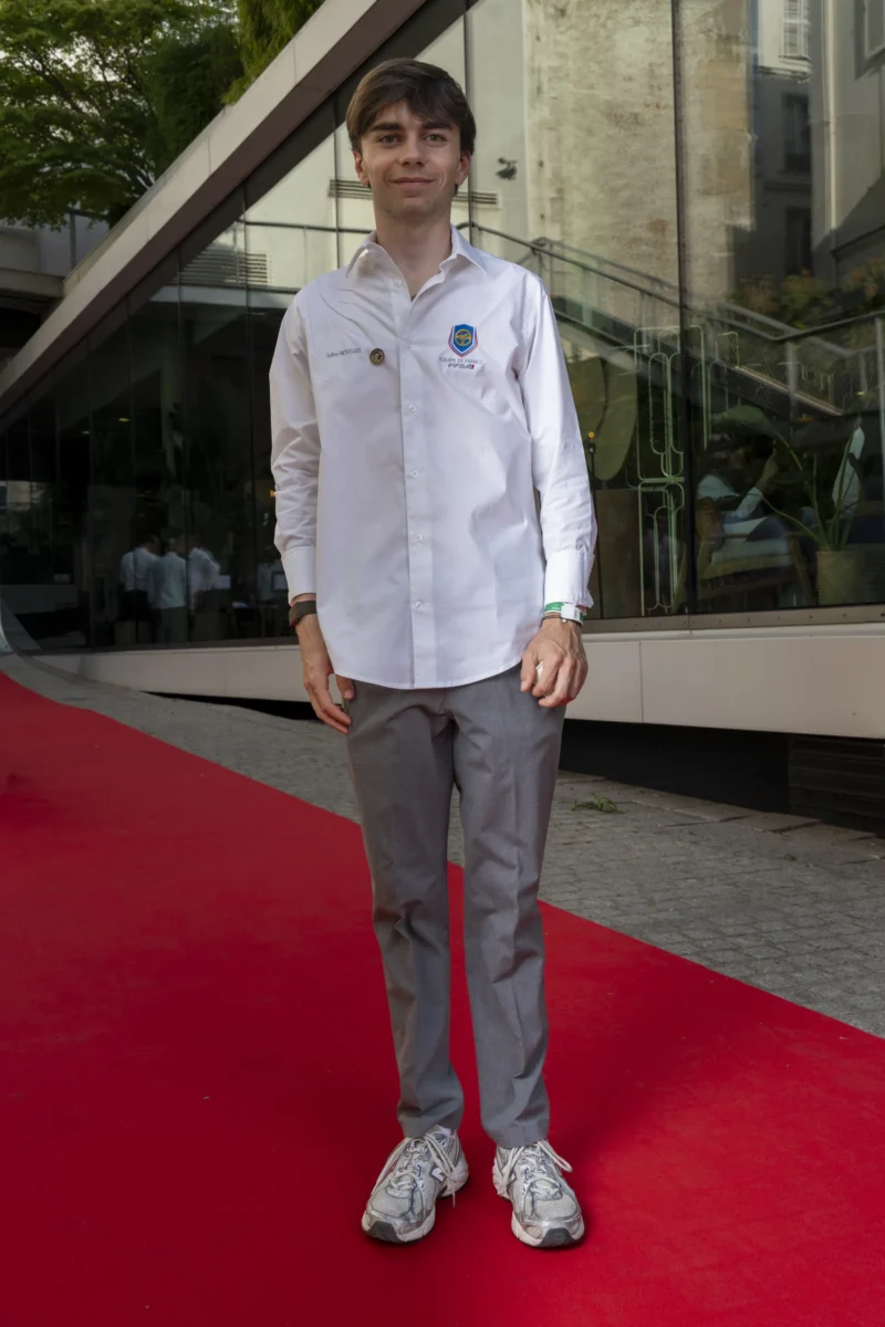 Cette photographie met en scène un jeune homme comme sujet principal, debout sur un tapis rouge. Il porte une tenue décontractée : un blouson blanc, un pantalon gris et des baskets.Le cadrage est serré, centré sur lui, avec un arrière-plan flou composé d’une structure architecturale moderne. La lumière semble naturelle, douce et légèrement latérale, créant des ombres discrètes. Les couleurs dominantes sont les tons neutres (gris, blanc) et le rouge vif du tapis.Le style est sobre et contemporain, évoquant une ambiance semi-formelle, peut-être liée à un événement public ou culturel.