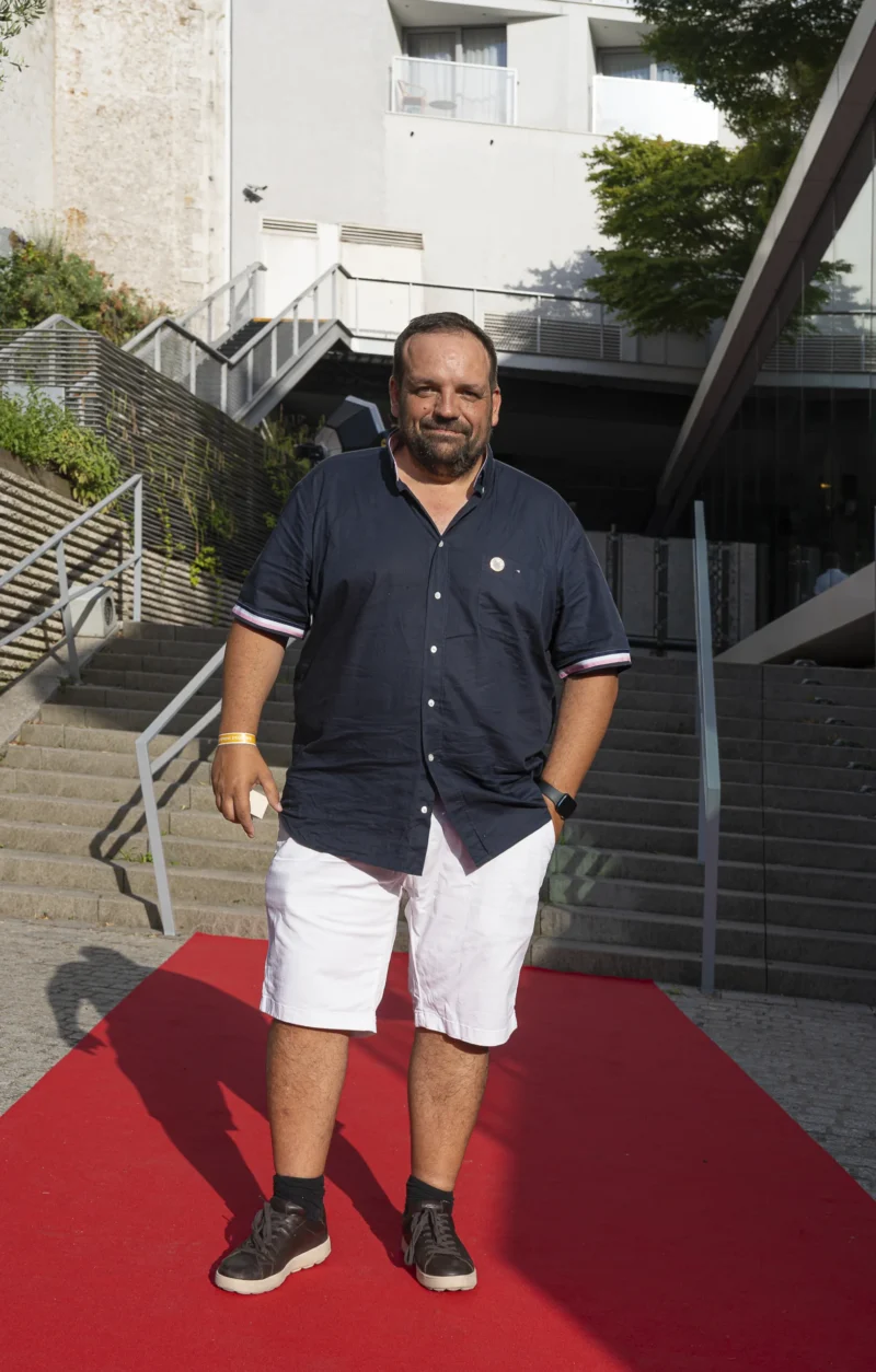 Cette photographie montre un homme comme sujet principal, debout sur un tapis rouge. Il porte une chemise bleue, un short blanc et des chaussures noires. La composition met en valeur les escaliers en pierre et une façade moderne en arrière-plan.Le cadrage est serré sur le sujet, avec une légère inclinaison vers le haut. La lumière semble naturelle, bien répartie, créant des ombres douces. Les couleurs dominantes sont les tons bleus, blancs et gris, avec une touche de rouge vif du tapis. L’ambiance est à la fois décontractée et légèrement formelle, évoquant un événement public ou une réception.