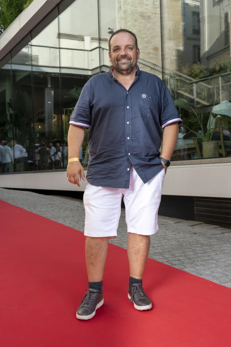 Cette photographie met en scène un homme comme sujet principal, debout sur un tapis rouge. Il porte un polo bleu clair et un short blanc, avec des chaussures noires.La composition est simple et centrée, avec un cadrage serré sur le sujet. L’arrière-plan montre une façade moderne avec des éléments architecturaux et des plantes.La lumière est naturelle et douce, éclairant bien le sujet. Les couleurs dominantes sont les tons bleus, blancs et gris, avec une touche de rouge due au tapis.L’ambiance est élégante et décontractée, évoquant un événement public ou une cérémonie informelle.