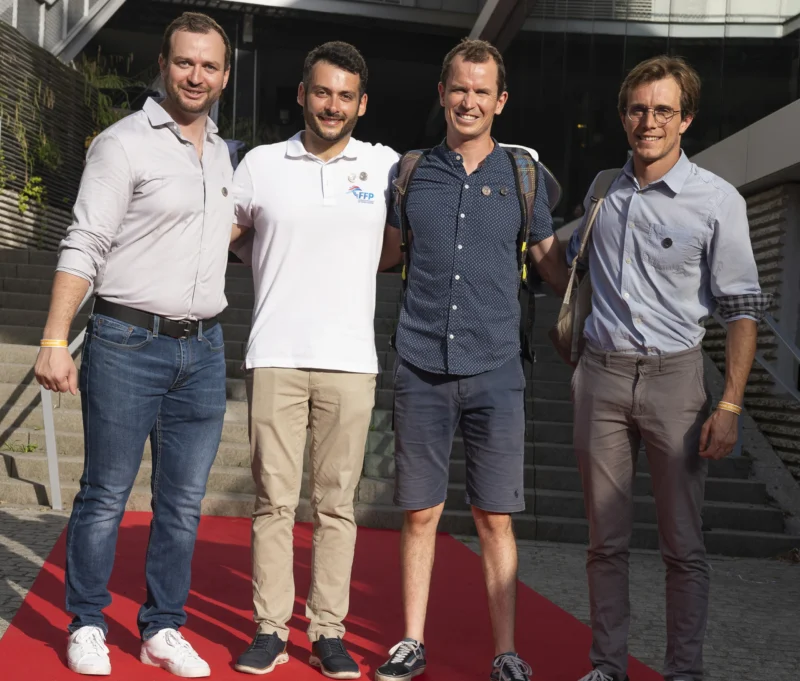 Cette photographie montre quatre hommes posant sur un tapis rouge. Le sujet principal est leur présence ensemble, suggérant un événement formel ou médiatique.La composition est symétrique, avec les individus alignés côte à côte. Le cadrage est serré, mettant en valeur leurs visages et leurs tenues décontractées mais soignées. La lumière est naturelle et douce, probablement en extérieur.Les couleurs dominantes sont neutres : gris, bleu clair et blanc, avec des touches de rouge sur le tapis. Le style est sobre et moderne, évoquant une ambiance professionnelle et détendue.
