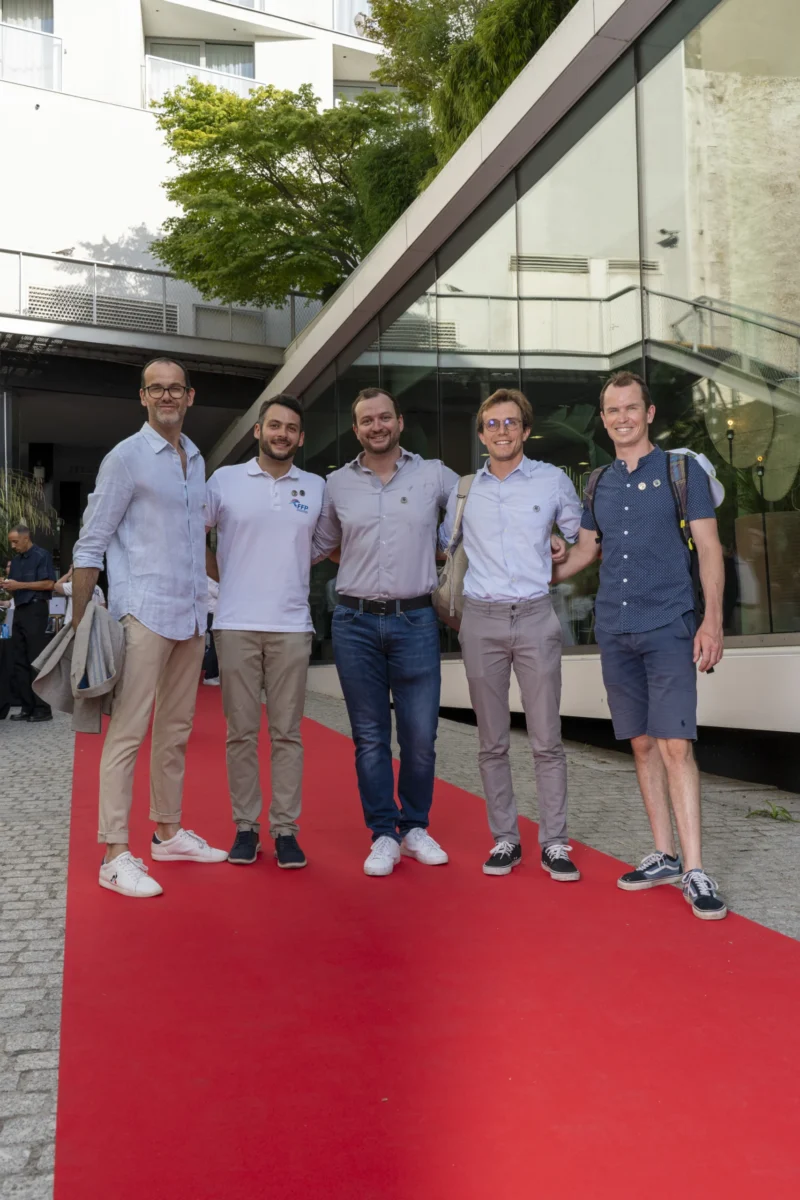 Cette photographie montre cinq hommes debout sur un tapis rouge. Le sujet principal est leur groupe, aligné et souriant, suggérant une ambiance conviviale ou professionnelle.La composition est symétrique, avec un cadrage serré sur les individus. L'arrière-plan, avec un bâtiment moderne et des arbres, est légèrement flou. La lumière est naturelle, douce et diffuse, probablement en journée. Les couleurs dominantes sont les tons neutres (gris, beige) et le rouge vif du tapis. Le style est sobre et contemporain, évoquant une ambiance formelle mais détendue.
