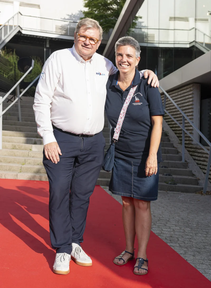 Cette photographie montre deux personnes posant sur un tapis rouge. Le sujet principal est un homme et une femme souriants, habillés de manière décontractée mais élégante.La composition est symétrique, avec les deux sujets côte à côte. Le cadrage est serré, mettant en valeur leurs visages et le haut de leur corps. La lumière est naturelle et douce, éclairant uniformément les sujets. Les couleurs dominantes sont les tons neutres (gris, blanc) de leurs vêtements, contrastant avec le rouge vif du tapis.L’ambiance est conviviale et professionnelle, évoquant un événement public ou une cérémonie.