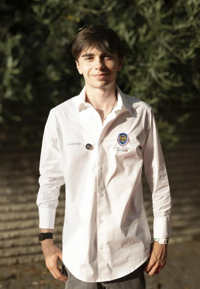 Cette photographie montre un jeune homme en tenue blanche, probablement un médecin ou un étudiant en médecine, identifiable par son badge et son stéthoscope. Le cadrage est serré sur lui, avec un arrière-plan flou de végétation.La lumière naturelle, douce et légèrement latérale, éclaire son visage et met en valeur les détails de sa tenue. Les couleurs dominantes sont les tons blancs et verts, créant une ambiance sobre et professionnelle. Le style est épuré et naturel, évoquant une scène authentique et calme.