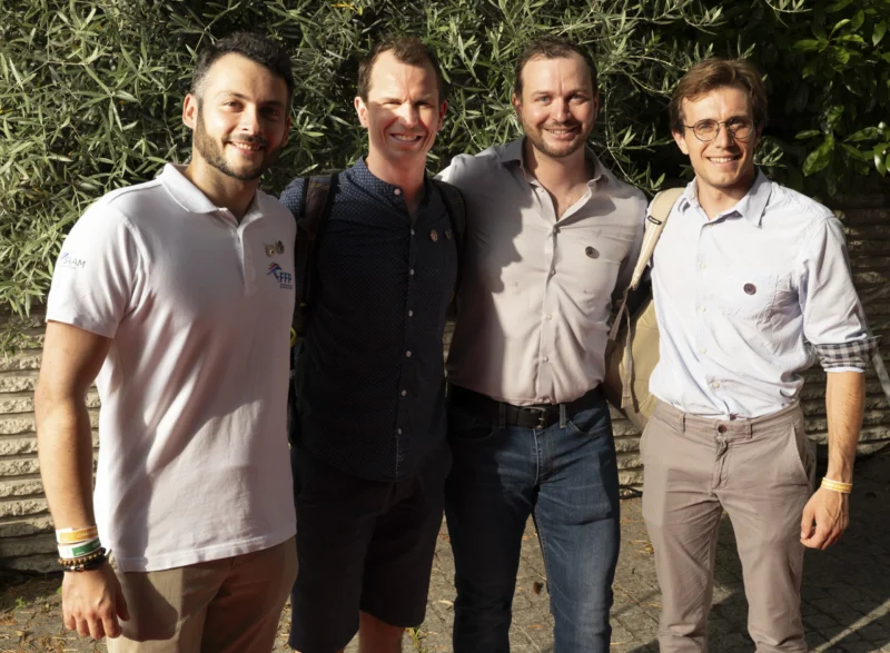 Cette photographie montre quatre hommes souriants posant côte à côte. Le sujet principal est leur interaction amicale.La composition est symétrique, avec un cadrage serré sur les épaules et le haut du corps. La lumière naturelle, douce et homogène, semble provenir d’une source externe, éclairant uniformément les visages.Les couleurs dominantes sont neutres : gris clair (vêtements), verts (arrière-plan) et touches de brun. Le style est simple et naturel, évoquant une ambiance détendue et conviviale, typique d’un moment informel entre amis.