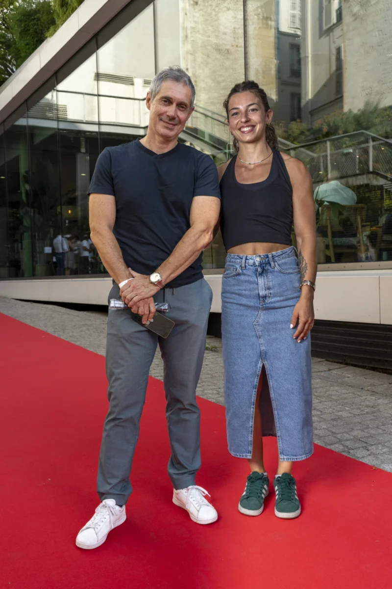 Cette photographie montre deux personnes posant sur un tapis rouge. Le sujet principal est un couple souriant, l’homme en haut noir et jeans, la femme en haut sport et jean déchiré avec des baskets colorées. La composition est symétrique, avec les deux sujets côte à côte. Le cadrage est serré, mettant en valeur leurs visages et leur tenue. La lumière est naturelle et douce, sans ombres marquées. Les couleurs dominantes sont les tons neutres (noir, gris) et les bleus des jeans, rehaussés par les touches de vert des baskets. L’ambiance est décontractée et joyeuse, évoquant un événement public ou une soirée.