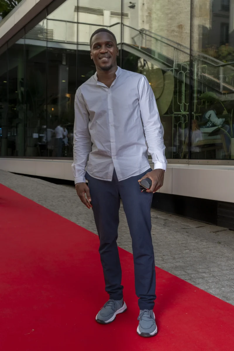 Cette photographie met en scène un jeune homme souriant, sujet principal, vêtu d’un pull blanc et d’un pantalon bleu, debout sur un tapis rouge. Le cadrage est serré, centré sur lui, avec un arrière-plan architectural moderne en verre et métal. La lumière est naturelle et douce, éclairant son visage sans ombres marquées. Les couleurs dominantes sont le blanc, le bleu et le rouge vif du tapis. Le style est épuré et contemporain, évoquant une ambiance formelle, peut-être un événement public ou une cérémonie. L’atmosphère est positive et élégante.