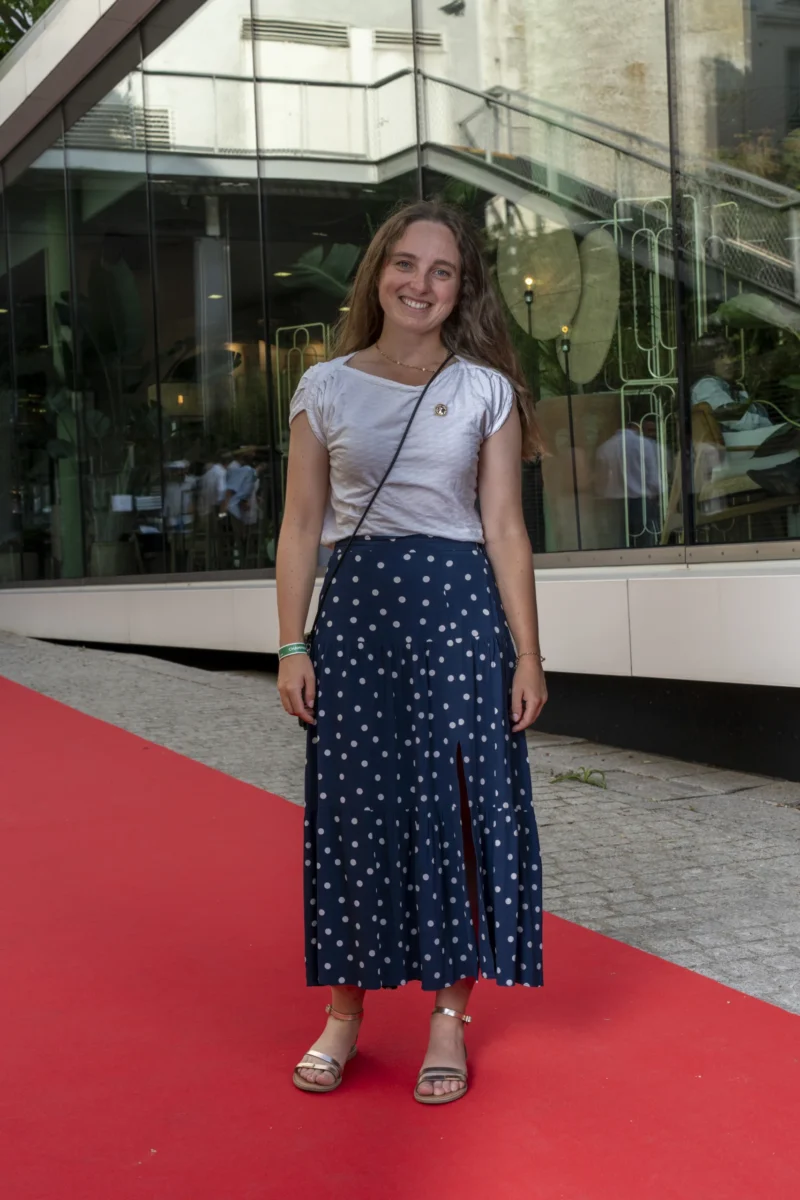 Cette photographie met en scène une jeune femme souriante, sujet principal, debout sur un tapis rouge. Elle porte une tenue décontractée : un haut blanc, une jupe à pois bleus et des espadrilles.Le cadrage est serré, centré sur elle, avec un arrière-plan flou montrant une façade moderne et des vitrines. La lumière est naturelle et douce, éclairant son visage sans ombre marquée. Les couleurs dominantes sont les bleus de la jupe, le blanc du haut, et le rouge du tapis, créant un contraste dynamique.L’ambiance est à la fois élégante et décontractée, évoquant un événement public ou une sortie informelle.