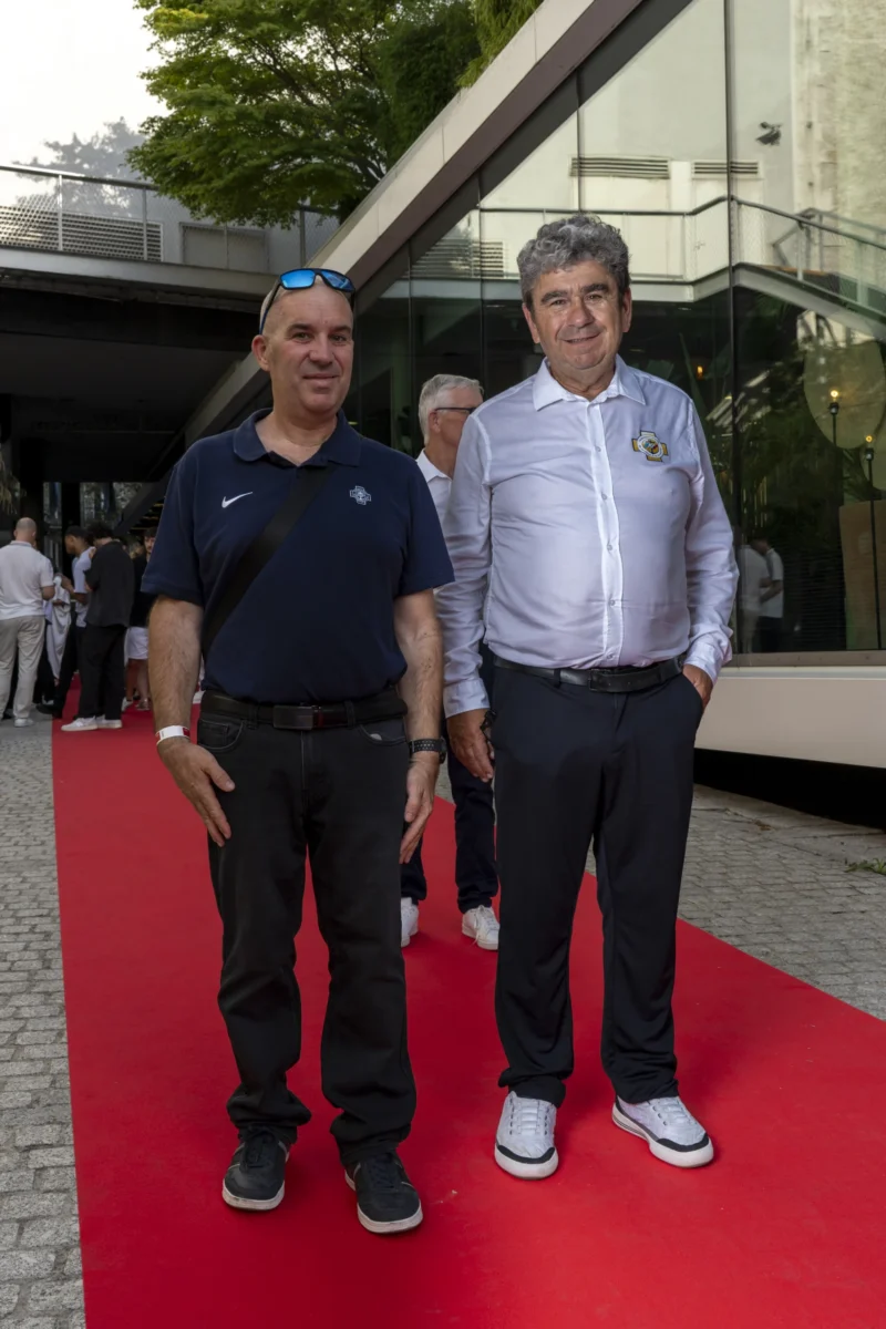 Cette photographie montre deux hommes posant sur un tapis rouge. Le sujet principal est ces deux individus, l’un en polo bleu et l’autre en chemise claire, souriants et détendus. La composition est symétrique, avec les deux hommes placés côte à côte au centre.Le cadrage est serré sur eux, avec un arrière-plan flou montrant une structure architecturale et des personnes. La lumière est naturelle et douce, sans ombres marquées. Les couleurs dominantes sont les tons neutres (gris, bleu) et le rouge vif du tapis. L’ambiance est conviviale et légèrement formelle, évoquant un événement public ou une cérémonie.