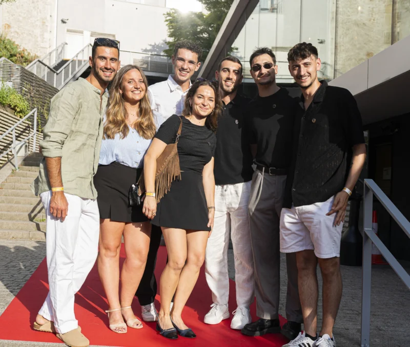 Cette photographie montre un groupe de six personnes posant sur un tapis rouge. Le sujet principal est l’ensemble du groupe, composé de trois hommes et trois femmes, habillés élégamment.La composition est symétrique, avec les individus alignés côte à côte. Le cadrage est serré, mettant en valeur leurs visages et tenues. La lumière est naturelle et douce, probablement en extérieur en journée. Les couleurs dominantes sont neutres (noir, blanc, beige) avec des touches de bleu et vert en arrière-plan.L’ambiance est festive et formelle, évoquant un événement mondain ou une cérémonie.