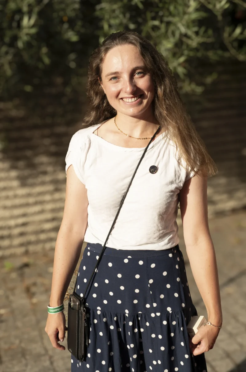 Cette photographie met en scène une jeune femme souriante, sujet principal, en position légèrement de trois-quarts. Elle porte une tenue estivale composée d’un haut blanc et d’une jupe à pois.Le cadrage est serré, centré sur elle, avec un arrière-plan flou de végétation et de murs, suggérant un extérieur naturel. La lumière est douce et naturelle, probablement en plein jour, éclairant son visage et créant des ombres légères.Les couleurs dominantes sont les tons clairs (blanc, beige) et les teintes pastel de la jupe. Le style est simple et spontané, évoquant une ambiance détendue et joyeuse.