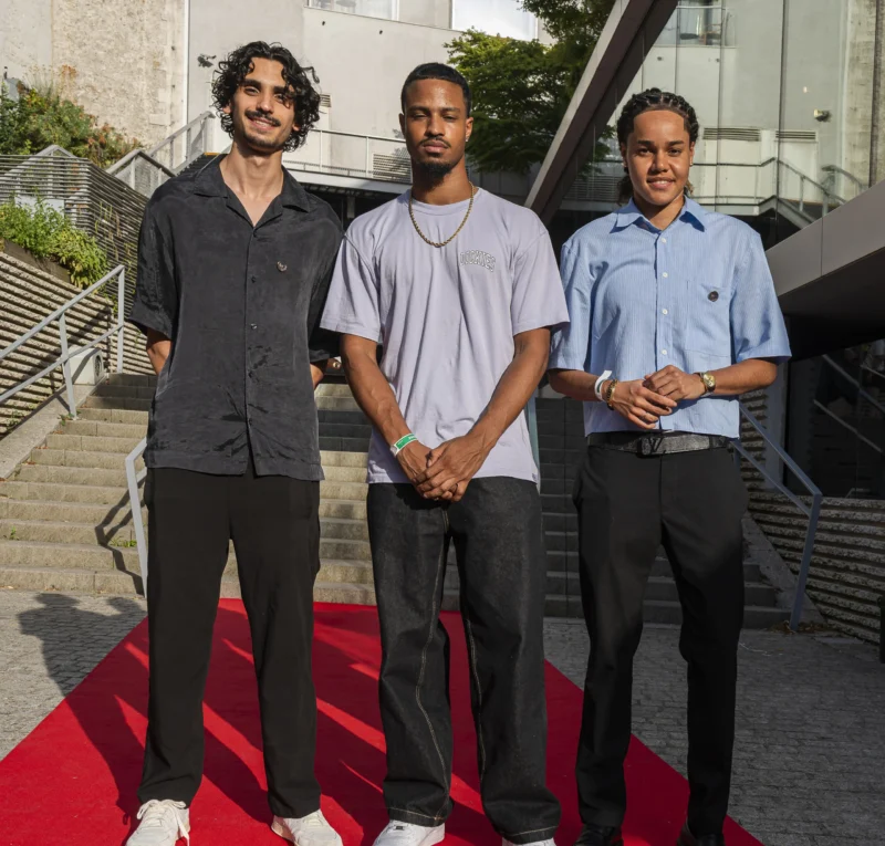 Cette photographie montre trois hommes posant côte à côte sur un tapis rouge. Le sujet principal est leur présence unie, avec une composition symétrique et un cadrage serré sur eux.La lumière est naturelle et douce, probablement en extérieur. Les couleurs dominantes sont les tons neutres (gris, bleu clair, noir) et le rouge vif du tapis. Le style est sobre et contemporain, évoquant une ambiance professionnelle ou événementielle, avec une touche d’élégance décontractée.
