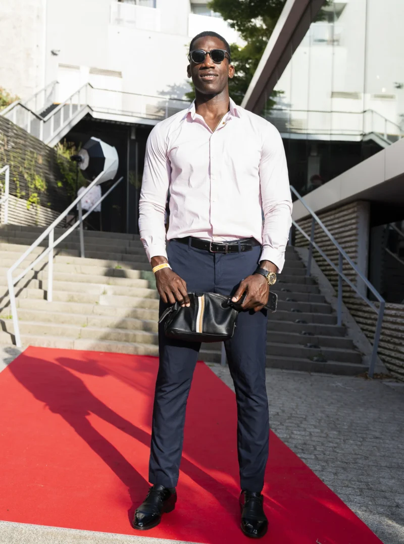 Cette photographie met en scène un homme comme sujet principal, vêtu d'un style élégant et décontracté : chemise blanche, pantalon noir et lunettes de soleil. Le cadrage est serré, le plaçant sur un tapis rouge, suggérant un événement formel.La lumière semble naturelle, avec des ombres douces, et l’ambiance est sophistiquée. Les couleurs dominantes sont les tons neutres (noir, blanc, gris) rehaussés par le rouge vif du tapis. L’arrière-plan architectural et les escaliers ajoutent une touche urbaine et moderne, renforçant un style contemporain et chic.