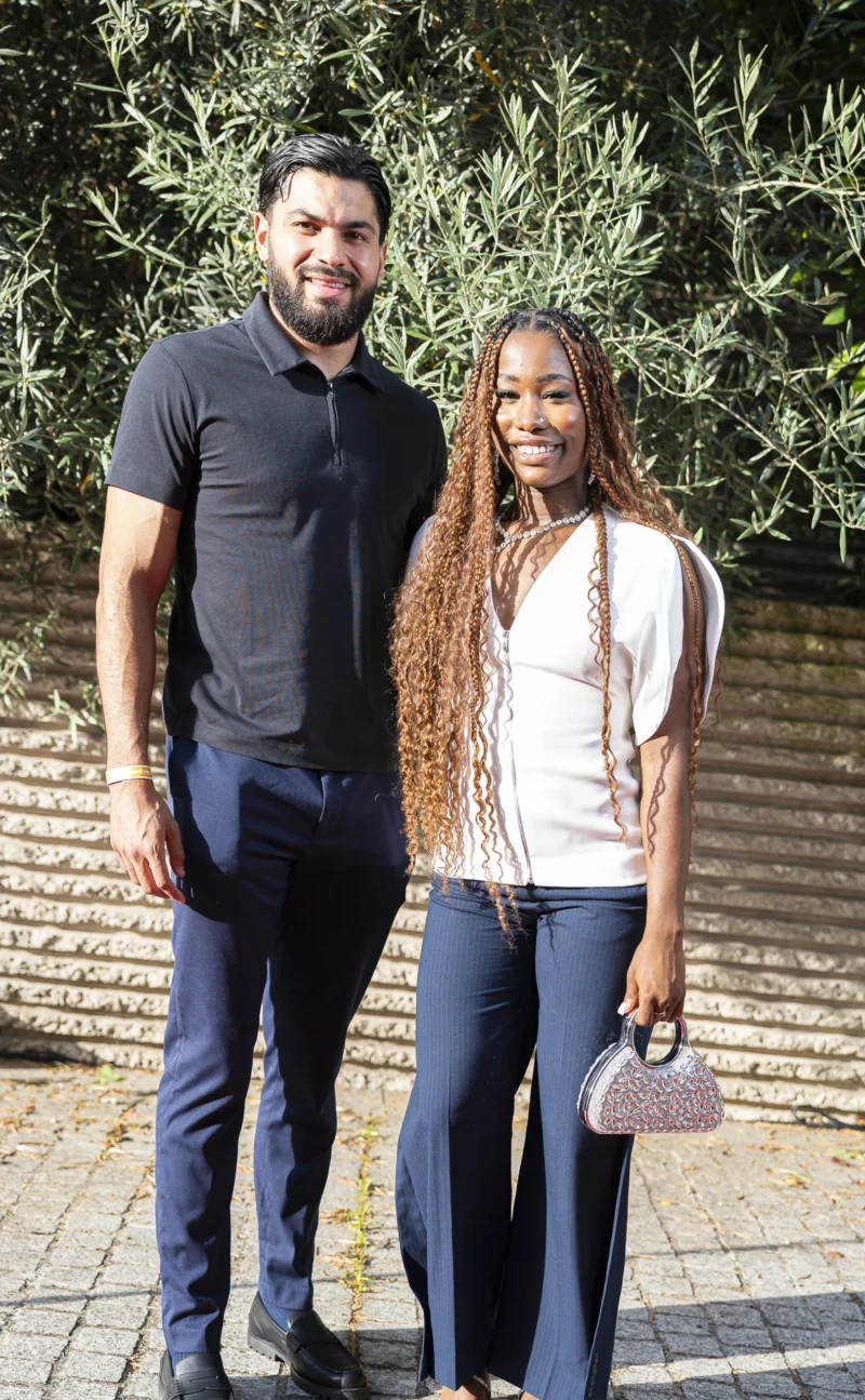 Cette photographie montre deux personnes posant côte à côte. Le sujet principal est un couple souriant, l’homme vêtu d’un polo noir et la femme en haut blanc avec un jean, tenant un accessoire rose.La composition est symétrique et équilibrée, avec un cadrage serré sur les épaules et le haut du corps. L’arrière-plan flou, avec des éléments végétaux, suggère un extérieur lumineux. Les couleurs dominantes sont les tons neutres (noir, blanc, bleu) rehaussés par les touches de rose et vert.Le style est naturel et décontracté, évoquant une ambiance chaleureuse et conviviale.