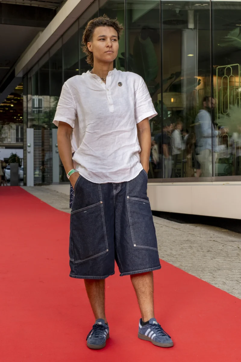 Cette photographie met en scène un homme comme sujet principal, vêtu d’un polo blanc et de shorts bleus, debout sur un tapis rouge. Le cadrage est serré, centré sur lui, avec un arrière-plan flou montrant une entrée de bâtiment et des personnes indistinctes. La lumière est naturelle et douce, éclairant le sujet de face. Les couleurs dominantes sont les tons bleus des shorts, le blanc du polo et le rouge vif du tapis. L’ambiance suggère un événement formel, comme une première ou un festival, avec une touche décontractée apportée par le style vestimentaire du sujet.