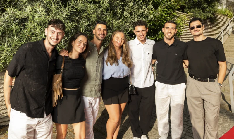 Cette photographie montre un groupe de six jeunes posant ensemble devant une végétation luxuriante. Le sujet principal est ce groupe uni, formant une composition harmonieuse en rang serré.Le cadrage est serré sur les personnes, avec un arrière-plan flou mettant en valeur les sujets. La lumière semble naturelle et douce, probablement en extérieur par temps clair. Les couleurs dominantes sont des tons neutres (noirs, blancs, gris) avec des touches de couleurs pastel (bleu clair, rose).Le style est décontracté et informel, évoquant une ambiance conviviale et détendue, typique d’un moment de loisir ou de vacances.