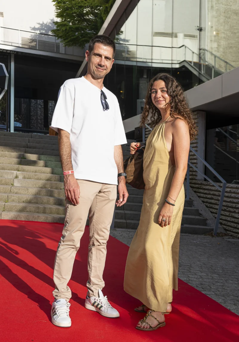 Cette photographie met en scène un couple sur un tapis rouge. Le sujet principal est un homme en tenue décontractée (t-shirt blanc, pantalon beige) et une femme en robe longue jaune pâle. La composition est symétrique, avec les deux sujets alignés verticalement. Le cadrage est serré sur eux, incluant partiellement un bâtiment moderne en arrière-plan.La lumière est naturelle et douce, éclairant clairement les visages. Les couleurs dominantes sont les tons neutres (gris du bâtiment) et chauds (jaune de la robe, beige des vêtements). L’ambiance est élégante et décontractée, évoquant un événement semi-formel.