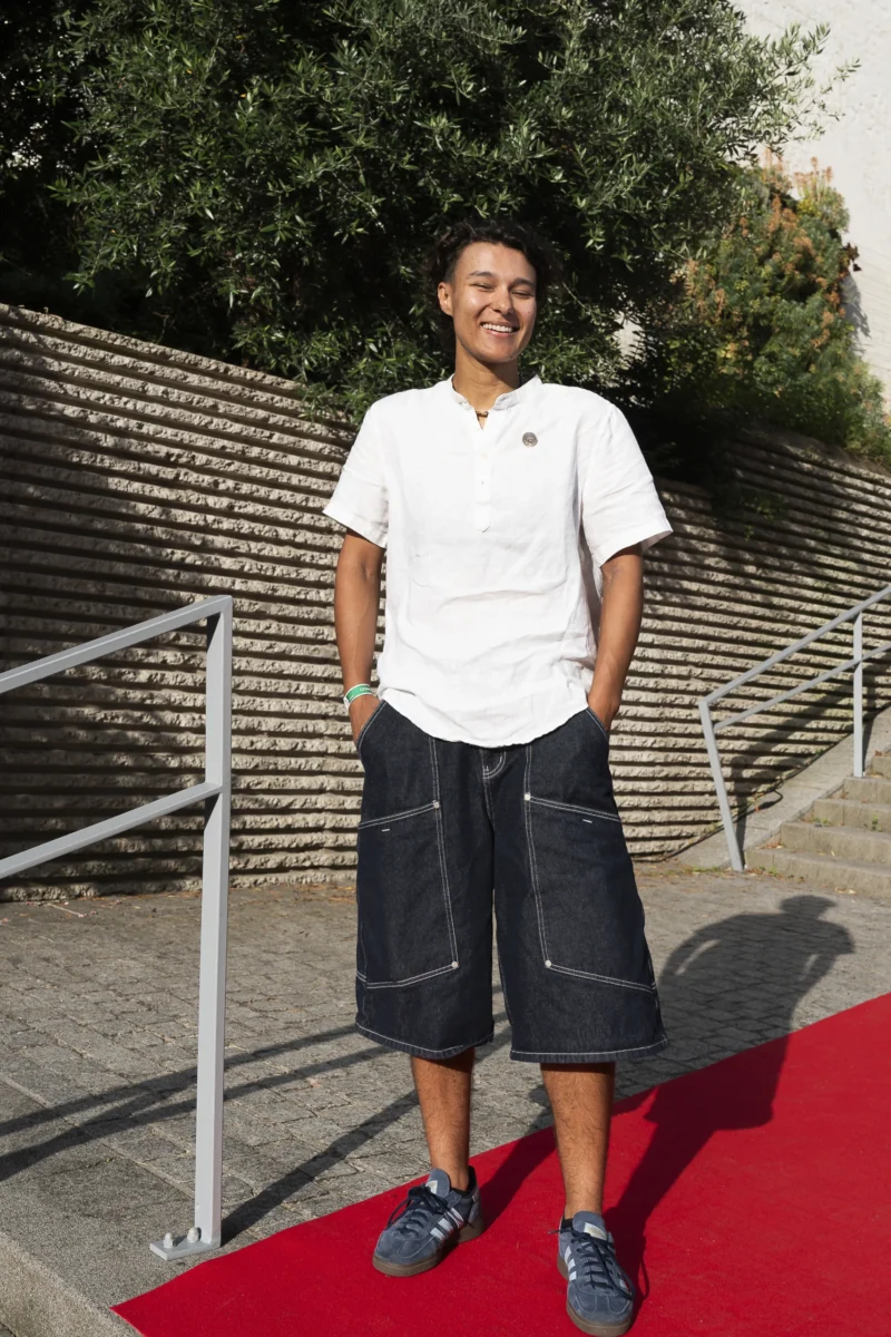 Cette photographie met en scène un jeune homme comme sujet principal, debout sur un tapis rouge. Il porte un polo blanc et un short noir, avec une posture décontractée.La composition est simple et centrée, avec une mise en valeur par le tapis rouge et une rampe métallique en arrière-plan. Le cadrage est serré, mettant l’accent sur le personnage.La lumière semble naturelle et douce, créant des ombres peu marquées. Les couleurs dominantes sont les tons clairs (blanc du polo) et les couleurs neutres (noir du short, gris des murs). L’ambiance est à la fois élégante et décontractée, évoquant un événement public ou une cérémonie.