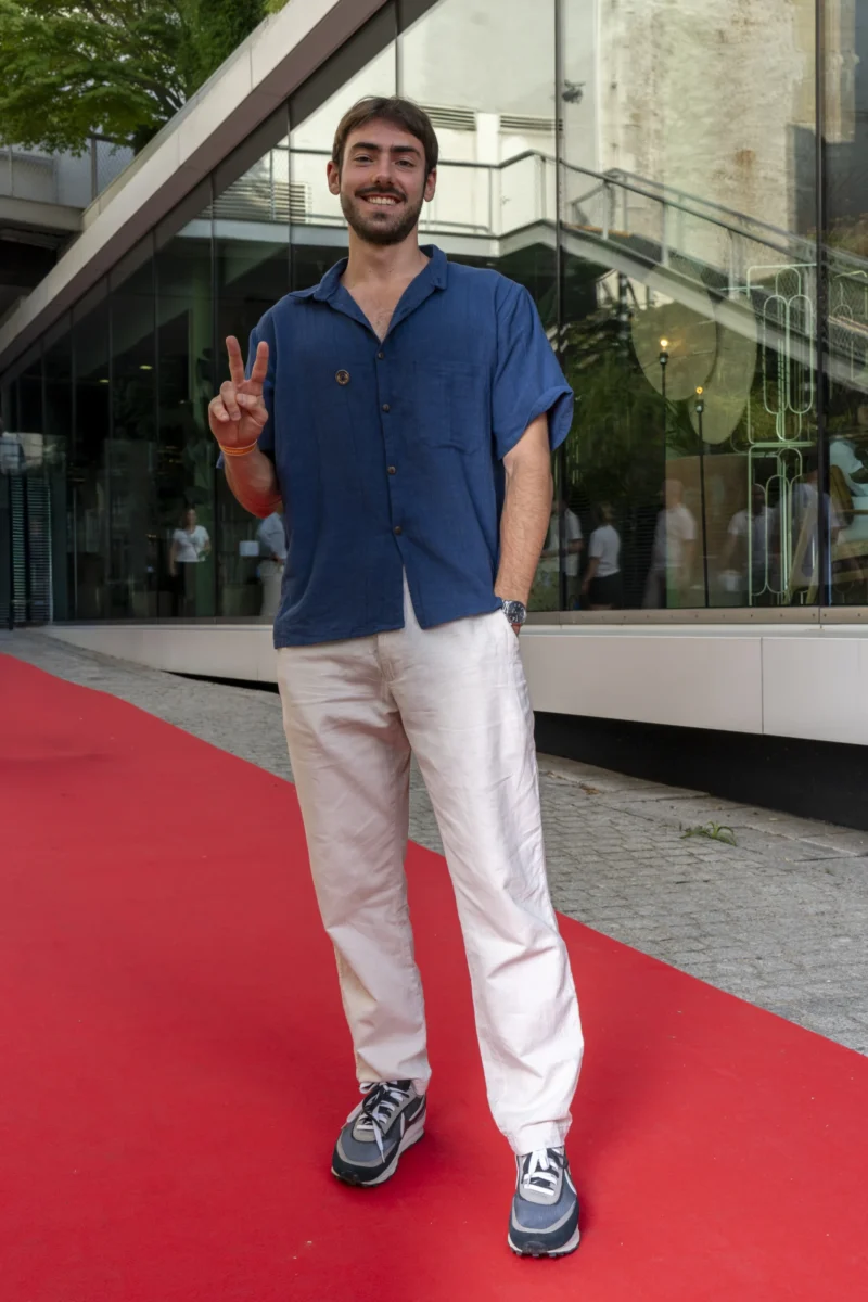 Cette photographie met en scène un homme comme sujet principal, debout sur un tapis rouge. Il porte un polo bleu et un pantalon blanc, avec des baskets, et fait un geste de paix.La composition est simple et centrée, avec un cadrage serré sur le sujet. L’arrière-plan montre une entrée moderne avec des colonnes et des vitres réfléchissantes. La lumière est naturelle et douce, éclairant bien le sujet.Les couleurs dominantes sont le bleu du vêtement, le blanc du pantalon, et le rouge du tapis. L’ambiance est décontractée et légèrement festive, évoquant un événement public ou une arrivée médiatique.