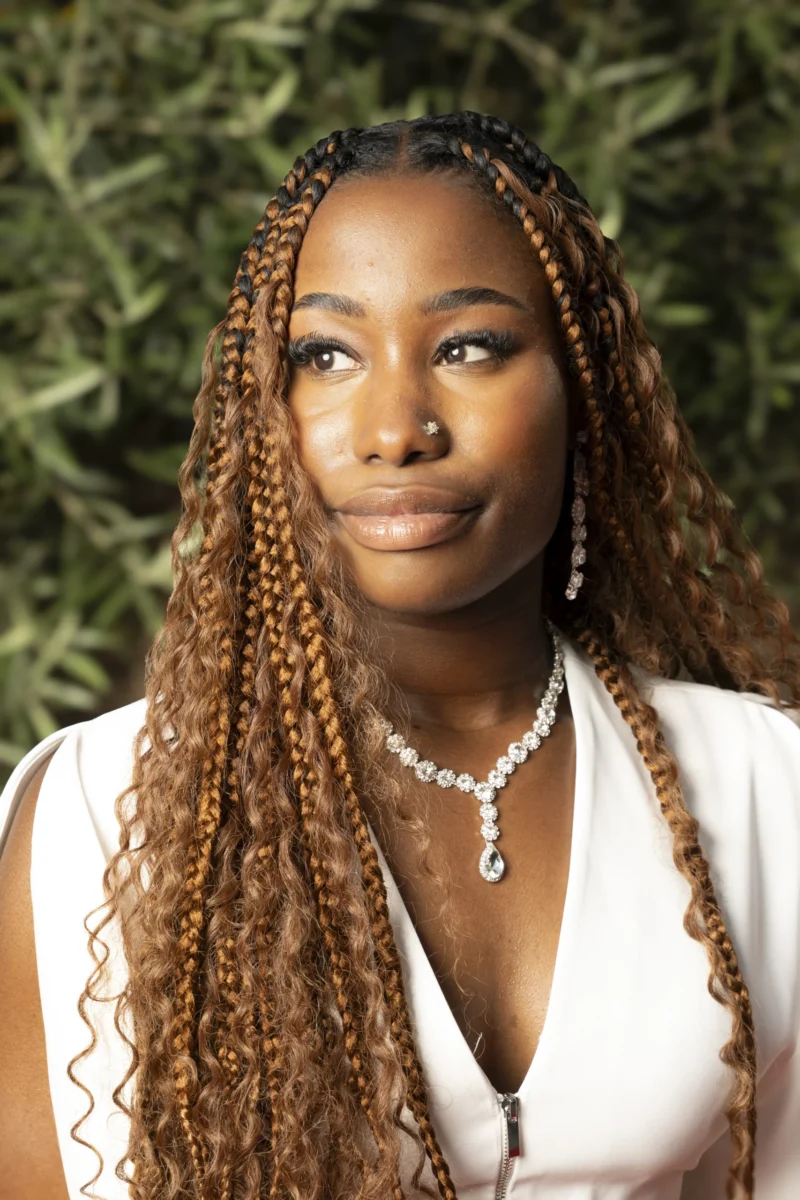 Cette photographie met en scène une femme au regard direct, sujet principal. Ses longs cheveux bouclés et son collier perlé attirent l'attention. La composition est simple et centrée, avec un cadrage serré sur son visage et ses épaules.La lumière est douce et naturelle, éclairant son visage sans ombres marquées. Les couleurs dominantes sont des tons chauds et neutres : blanc (vêtement), vert foncé (arrière-plan flou), et teints de peau naturels.Le style est épuré et élégant, évoquant une ambiance à la fois moderne et intemporelle.