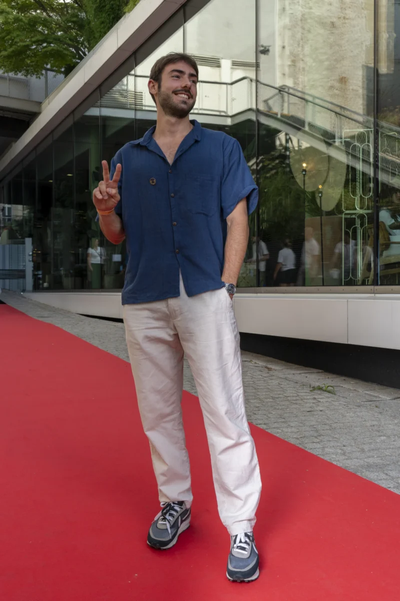 Cette photographie met en scène un homme comme sujet principal, vêtu d'une chemise bleue et d'un pantalon blanc, posant sur un tapis rouge. Le cadrage est serré, centré sur lui, avec un arrière-plan flou composé d'une structure en verre et de végétation.La lumière semble naturelle, bien répartie, mettant en valeur les tons clairs de ses vêtements. Les couleurs dominantes sont les bleus de sa chemise, les blancs de son pantalon, et le rouge vif du tapis. L'ambiance est décontractée mais élégante, évoquant un événement public ou une cérémonie. Le style est simple et moderne.