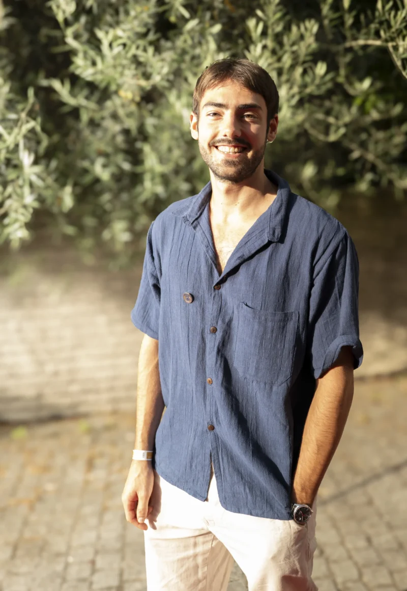 Cette photographie met en scène un jeune homme souriant, sujet principal, vêtu d'une chemise bleue et d'un pantalon clair. Le cadrage est serré, centré sur lui, avec un arrière-plan flou de végétation (feuillage vert).La lumière naturelle, douce et légèrement latérale, éclaire son visage, créant des ombres subtiles. Les couleurs dominantes sont les tons bleus de la chemise, les verts de la végétation et les clairs du pantalon et de la peau.Le style est naturel et décontracté, évoquant une ambiance détendue et chaleureuse, presque estivale.