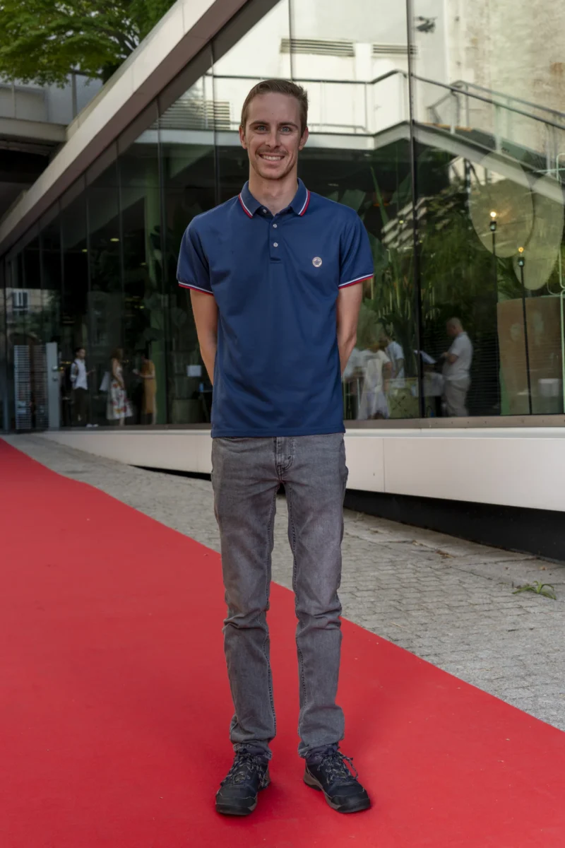 Cette photographie montre un homme en polo bleu et pantalon gris, debout sur un tapis rouge. Il est le sujet principal, positionné au centre du cadre. La composition est simple, avec un arrière-plan architectural moderne et des reflets sur une surface vitrée. Le cadrage est serré sur le sujet, mettant en valeur sa silhouette.La lumière semble naturelle, bien répartie, sans ombres marquées. Les couleurs dominantes sont le bleu du polo, le gris du pantalon, et le rouge vif du tapis. L’ambiance est sobre et professionnelle, évoquant un événement formel ou une inauguration.