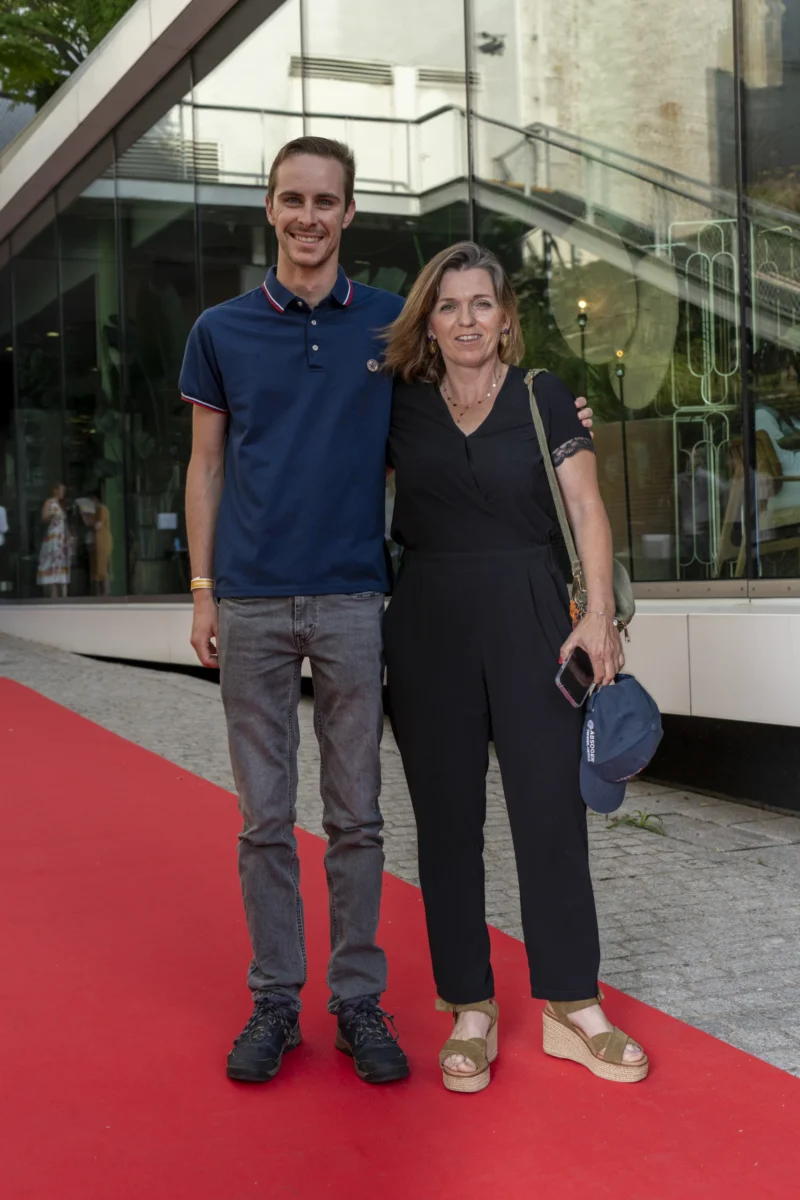 Cette photographie montre deux personnes posant sur un tapis rouge. Le sujet principal est un homme et une femme souriants, habillés de manière décontractée mais élégante.La composition est symétrique, avec les deux sujets côte à côte. Le cadrage est serré, mettant en valeur leurs visages et le haut de leur corps. La lumière est naturelle et douce, probablement en extérieur en journée. Les couleurs dominantes sont les tons neutres (gris, bleu marine, noir) et le rouge vif du tapis.Le style est simple et spontané, évoquant une ambiance conviviale et informelle, malgré le contexte formel du tapis rouge.