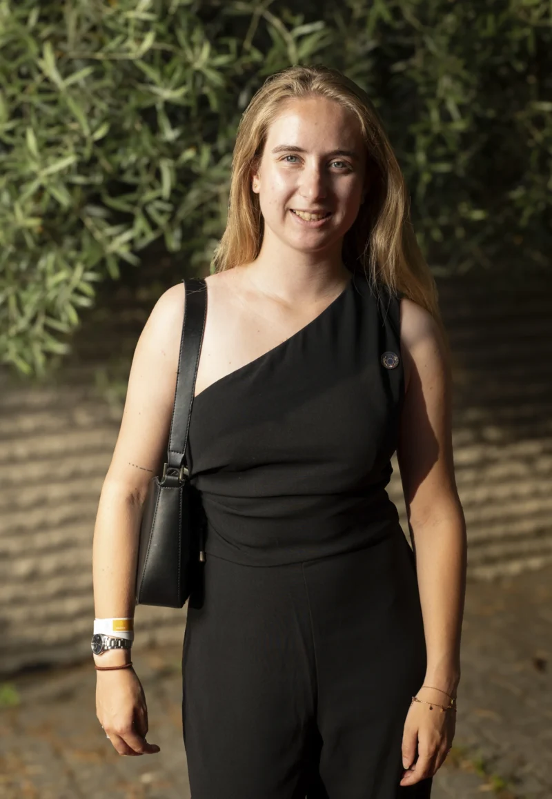 Cette photographie met en scène une jeune femme comme sujet principal. Elle porte une tenue noire élégante et accessoirisée simplement. Le cadrage est serré, centré sur elle, avec un arrière-plan flou de végétation, ce qui isole le sujet.La lumière semble naturelle et douce, probablement en extérieur. Les couleurs dominantes sont les tons sombres (noir) et les verts naturels de la végétation. Le style est sobre et contemporain, évoquant une ambiance détendue et professionnelle. L'expression souriante de la femme ajoute une touche chaleureuse.