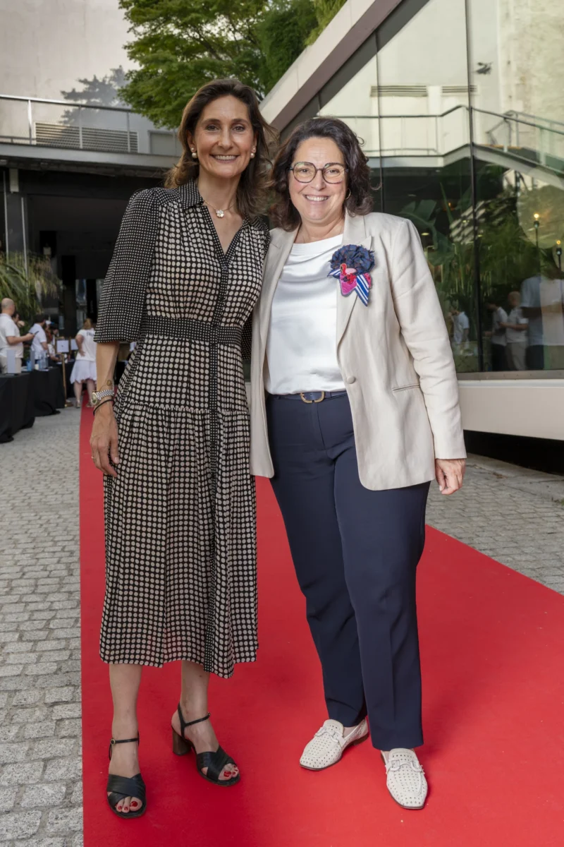 Cette photographie montre deux femmes posant côte à côte sur un tapis rouge. Le sujet principal est leur présence harmonieuse, l’une vêtue d’une robe à carreaux et l’autre d’un ensemble décontracté avec un pull et un badge.La composition est symétrique, avec un cadrage serré sur elles. La lumière est naturelle et douce, mettant en valeur leurs visages. Les couleurs dominantes sont les tons neutres (gris, bleu) et le rouge vif du tapis, créant un contraste élégant.L’ambiance est sophistiquée et conviviale, évoquant un événement formel ou culturel. Le style est à la fois chic et décontracté.
