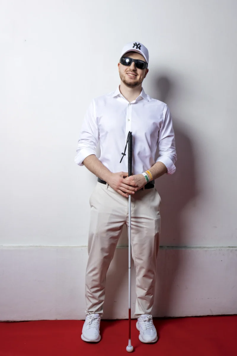 Cette photographie met en scène un homme comme sujet principal, vêtu d’un uniforme de caddie (pantalon beige, chemise blanche) et coiffé d’une casquette avec des lunettes. Il tient un drapeau de golf et pose sur un fond uni.Le cadrage est serré, centré sur le sujet, avec une composition symétrique. La lumière est douce et uniforme, éclairant le sujet sans ombres marquées. Les couleurs dominantes sont le blanc (chemise), le beige (pantalon) et le rouge (sol), créant un contraste visuel.Le style est épuré et professionnel, évoquant une ambiance à la fois classique et légèrement humoristique.