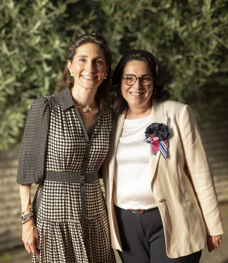 Cette photographie met en scène deux femmes souriantes posant côte à côte. Le sujet principal est leur complicité et leur élégance.La composition est symétrique, avec un cadrage serré sur elles. L'arrière-plan flou, composé de feuillage, isole les sujets. La lumière naturelle, douce et légèrement latérale, adoucit les traits et crée des ombres subtiles.Les couleurs dominantes sont neutres : gris, beige et blanc, rehaussées par une touche de bleu et de violet sur les accessoires. Le style est sobre et élégant, évoquant une ambiance chaleureuse et professionnelle.