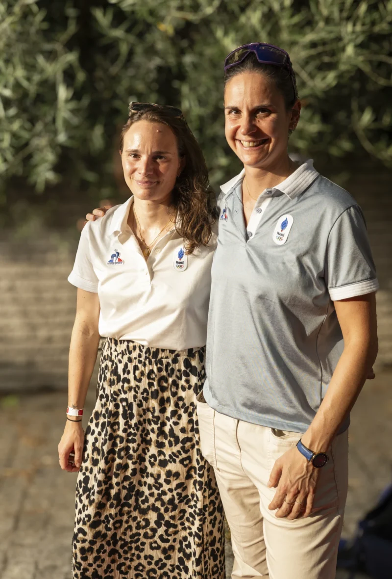 Cette photographie montre deux personnes souriantes posant côte à côte. Le sujet principal est ce duo, vêtu de tenues sportives (polo et jupe à motifs animaux). La composition est symétrique, avec un cadrage serré sur les épaules et le haut du corps.La lumière est naturelle et douce, probablement en extérieur. Les couleurs dominantes sont les tons neutres (gris du polo, beige de la jupe) avec des touches de vert en arrière-plan. L’ambiance est décontractée et chaleureuse, évoquant un moment convivial ou sportif.
