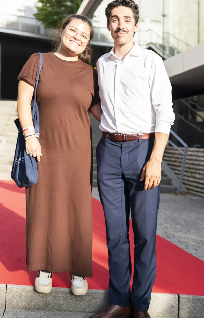 Cette photographie montre deux personnes posant côte à côte sur un tapis rouge. Le sujet principal est ce couple souriant, habillé élégamment : la femme porte une robe longue marron et des baskets, tandis que l’homme arbore une chemise blanche et un pantalon bleu avec une ceinture.Le cadrage est serré, centré sur eux. La lumière semble naturelle, douce et légèrement tamisée, créant une ambiance chaleureuse. Les couleurs dominantes sont les tons neutres (beige, marron, bleu clair, blanc), évoquant un style décontracté mais soigné. L’ambiance est conviviale et détendue, malgré le contexte formel suggéré par le tapis rouge.