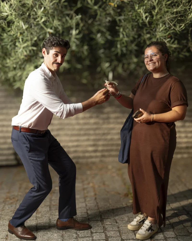 Cette photographie capture deux personnes en train de danser ou de partager un moment joyeux. Le sujet principal est leur interaction dynamique et souriante.La composition est équilibrée, avec un cadrage serré qui met en valeur leurs expressions et mouvements. La lumière naturelle, douce et légèrement latérale, adoucit les contours et crée une ambiance chaleureuse. Les couleurs dominantes sont les tons neutres (beige, marron) et les touches de vert des arbres en arrière-plan.Le style est naturel et spontané, évoquant une ambiance conviviale et détendue.