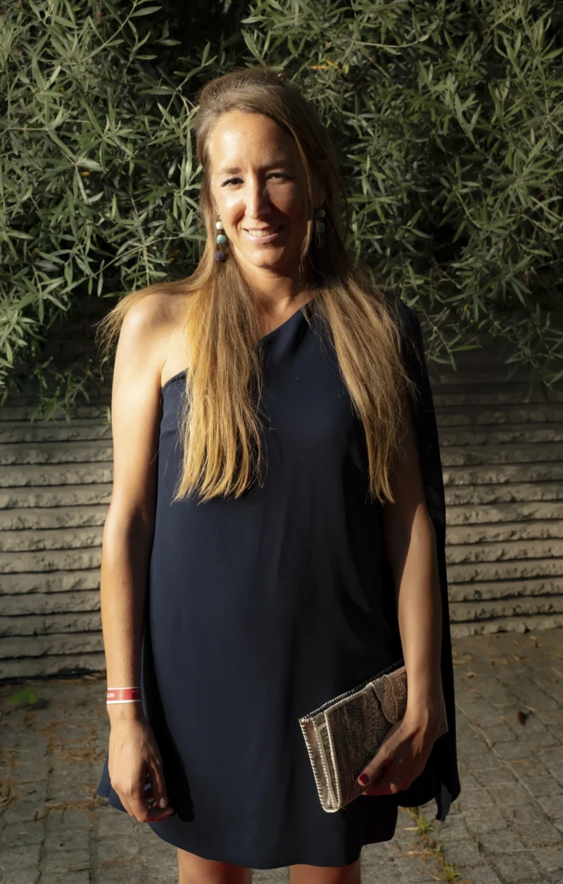 Cette photographie met en scène une femme souriante, sujet principal, vêtue d'une robe noire à manches courtes. Le cadrage est serré, centré sur elle, avec un arrière-plan flou de haies et de murs.La lumière naturelle éclaire son visage, créant des contrastes doux. Les couleurs dominantes sont les tons bleus-gris de sa robe et les verts naturels de la végétation. Le style est sobre et élégant, évoquant une ambiance détendue et chaleureuse, presque printanière.