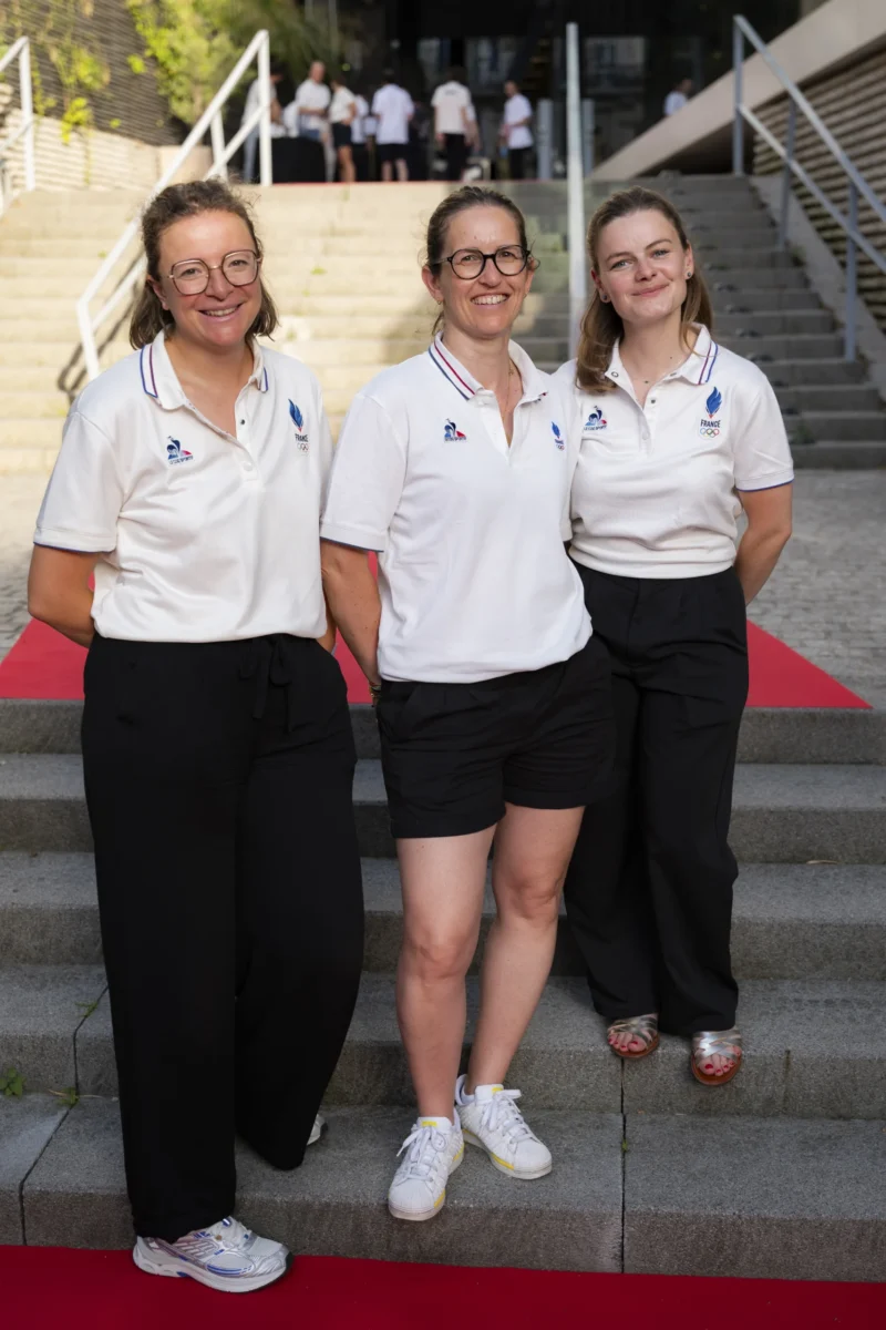 Cette photographie montre trois femmes posant ensemble sur un tapis rouge. Elles portent des tenues blanches identiques avec des logos, suggérant une appartenance à une même organisation ou équipe.Le cadrage est serré sur elles, avec un arrière-plan de marches en béton. La lumière est naturelle et douce, éclairant bien les visages. Les couleurs dominantes sont le blanc des vêtements, le rouge du tapis et les tons neutres de l’environnement.L’ambiance est professionnelle et formelle, avec une touche de solennité. Le style est sobre et direct.