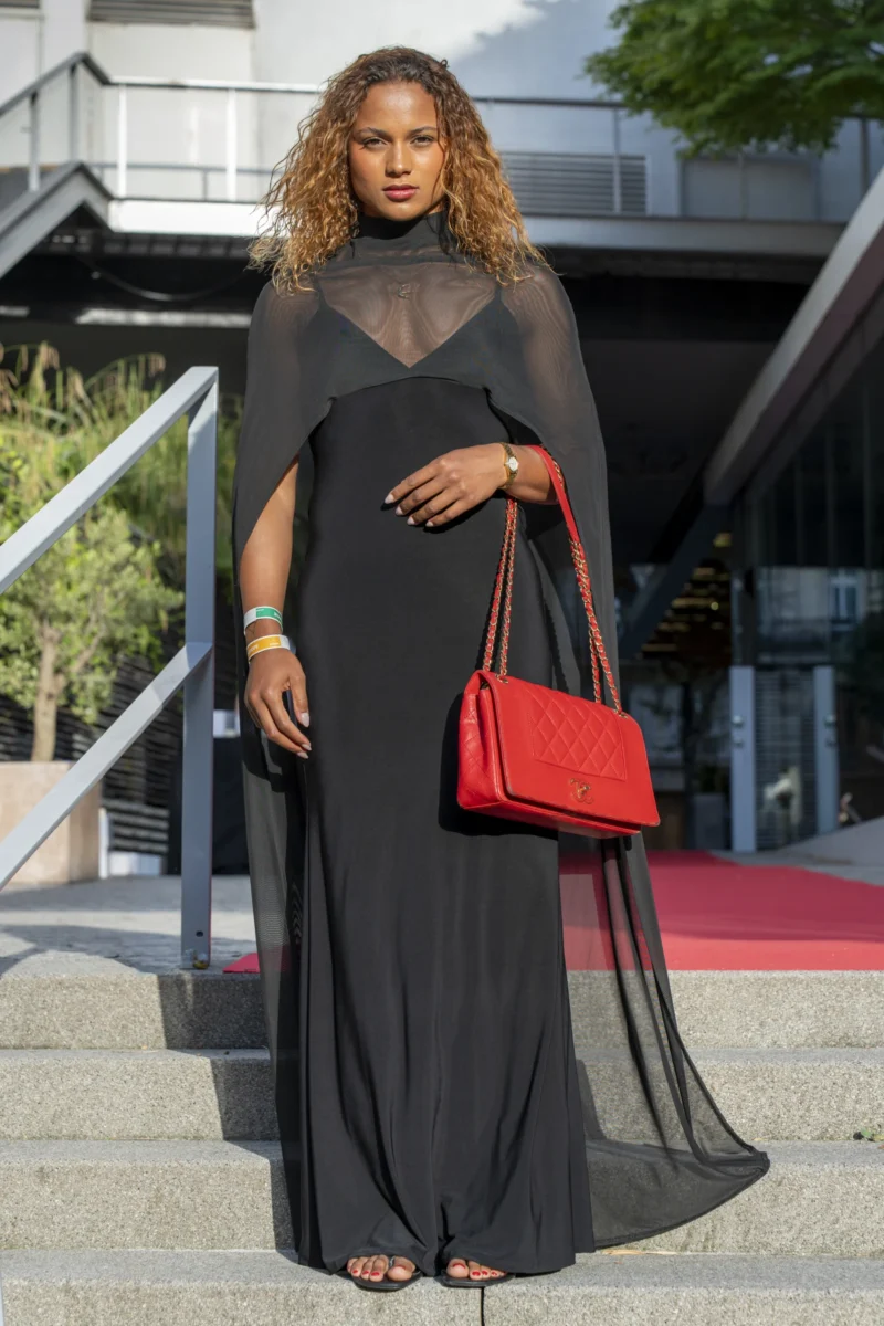 Cette photographie met en scène une femme comme sujet principal, vêtue d’une longue robe noire fluide et d’un manteau transparent assorti. Elle porte une ceinture rouge et un sac à main rouge vif.La composition est simple et élégante, avec un cadrage serré sur le sujet, mettant en valeur sa silhouette. L’arrière-plan, légèrement flou, montre des escaliers et une structure architecturale moderne.La lumière semble naturelle, douce et légèrement latérale, créant des jeux d’ombres et de clarté. Les couleurs dominantes sont le noir et le rouge, apportant un contraste moderne et sophistiqué. L’ambiance est chic et intemporelle.